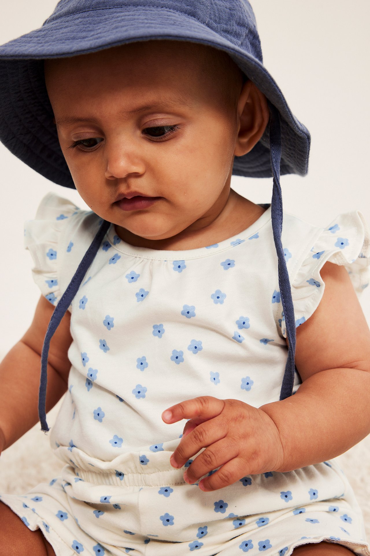 Nærbilde av baby i kremhvit blomstret body, volangermer og en blå bøttehatt.