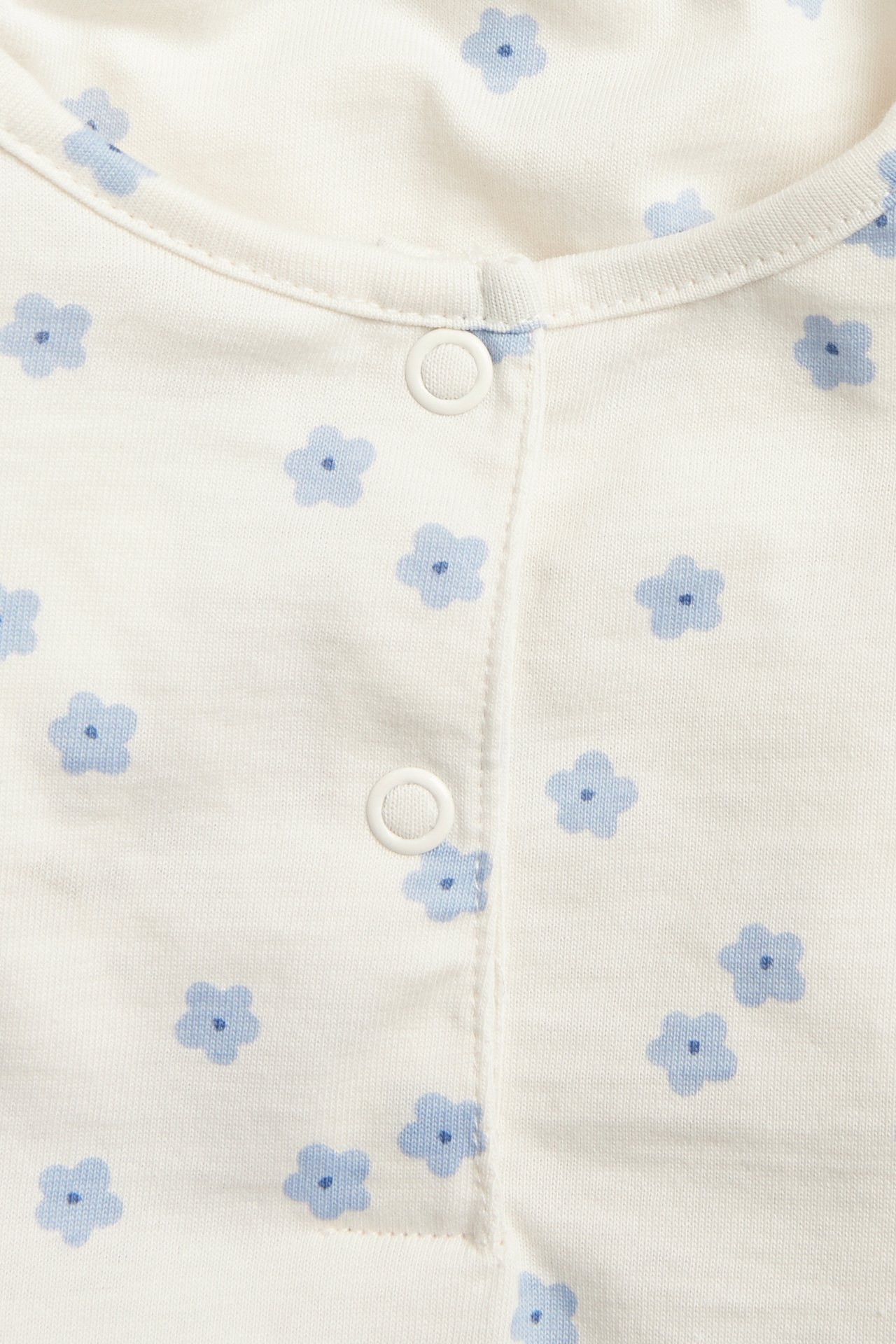 Nærbilde av en offwhite babybody i bomull med lyseblått blomstermønster og hvite trykknapper.