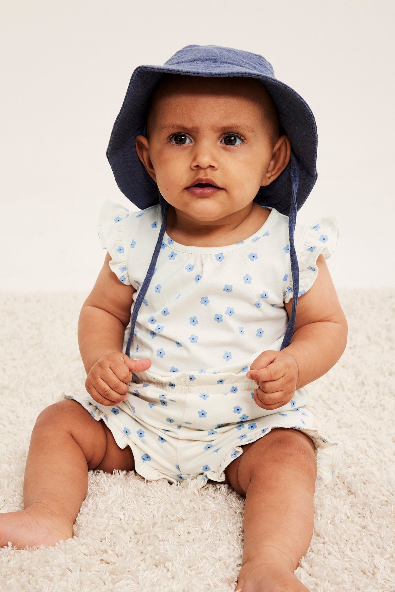 Forfra, baby i offwhite blomstret body med volangermer og marineblå bøttehatt.