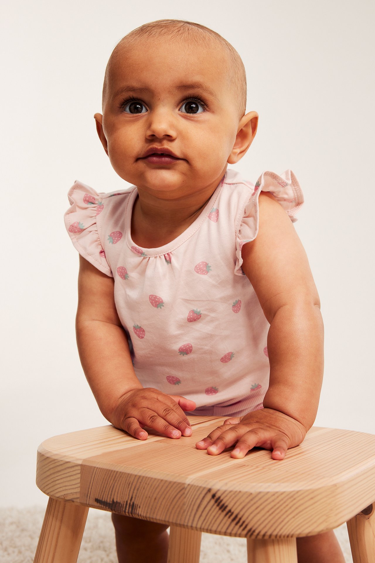 Edestä: Vauva yllään vaaleanpunainen puuvillabody, jossa röyhelöhihat ja mansikkakuvio.