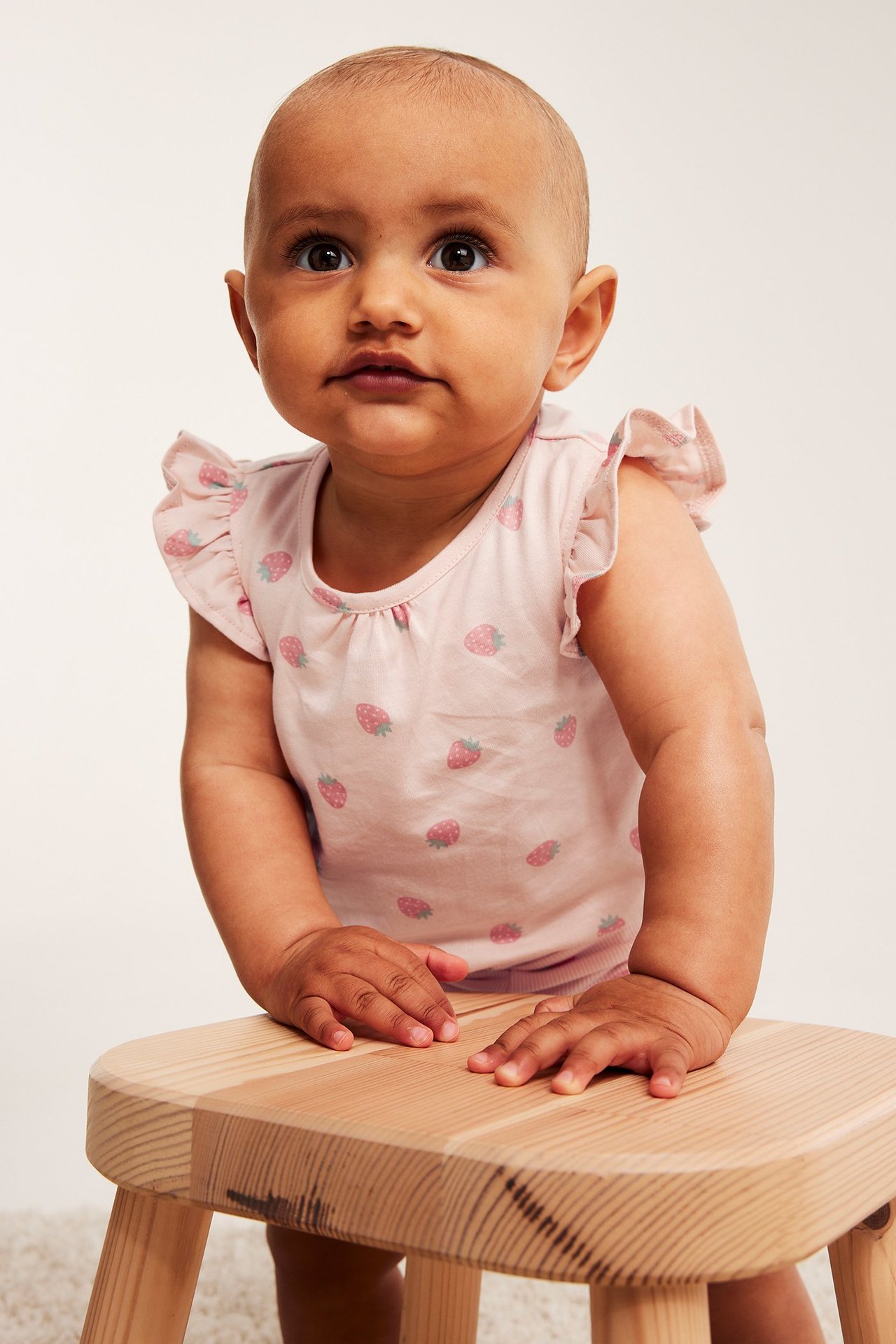 Widok z przodu: Niemowlę w jasnoróżowym bawełnianym body z falbankami i wzorem truskawek.