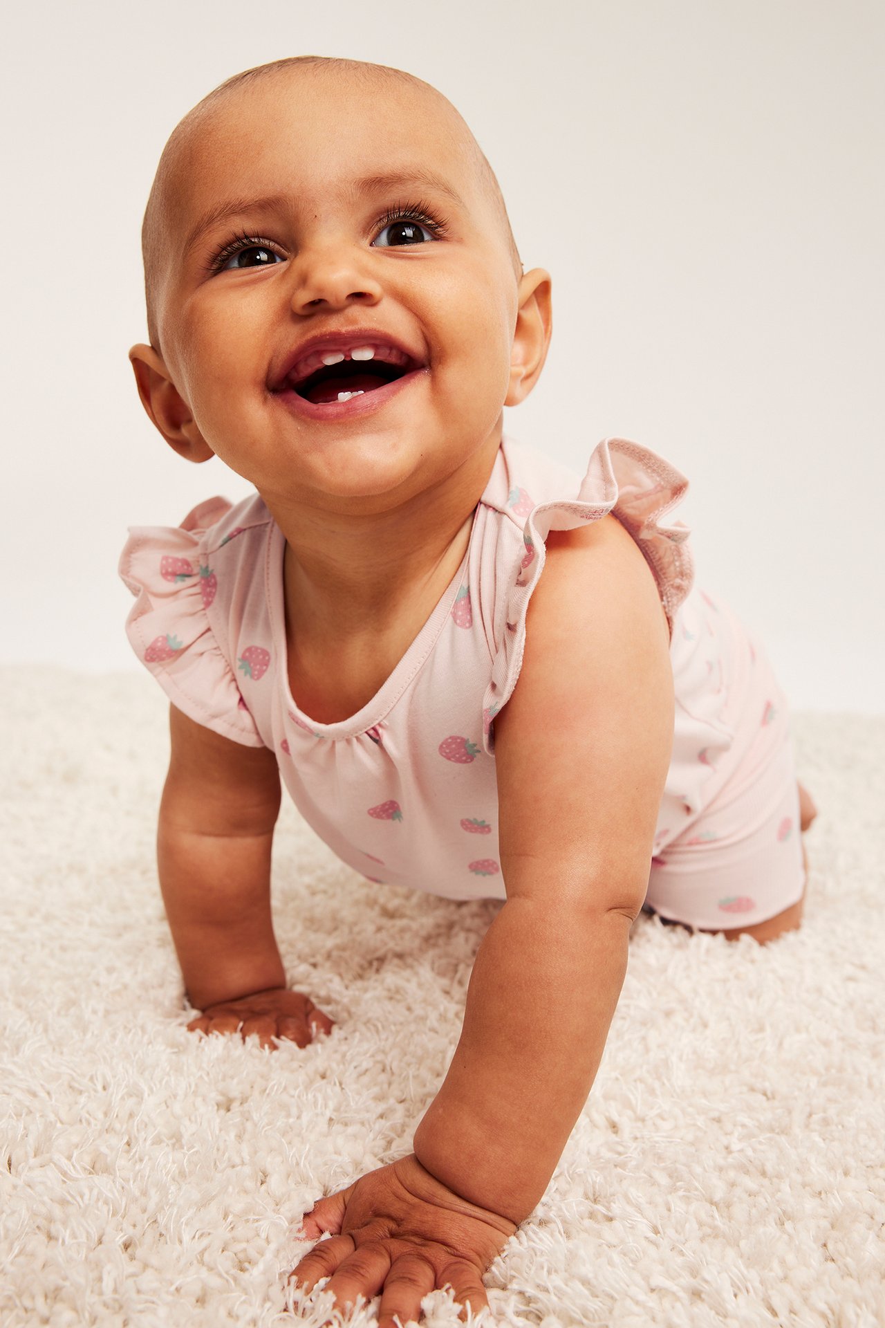 Edestä: Onnellinen vauva konttaa yllään vaaleanpunainen puuvillabody mansikkakuviolla ja röyhelöhihoilla.