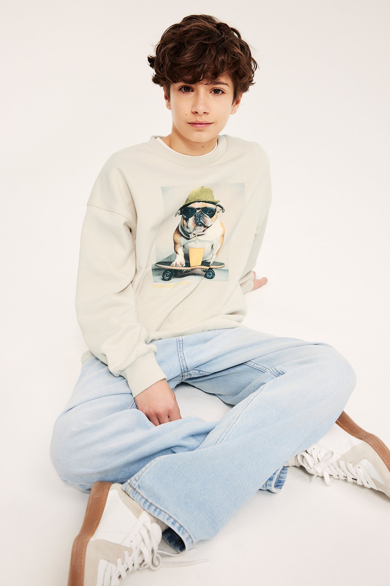Frontvisning: barn i lys beige bomullssweatshirt med bulldogtrykk, lyseblå vide jeans og hvite sneakers.