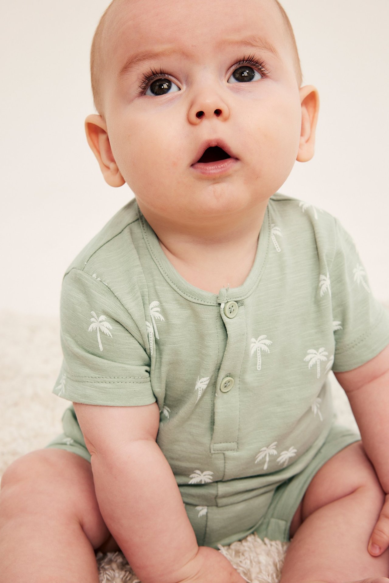 Częściowy widok z przodu niemowlęcia w jasnozielonym bawełnianym pajacyku z białym nadrukiem palm i listwą guzikową.
