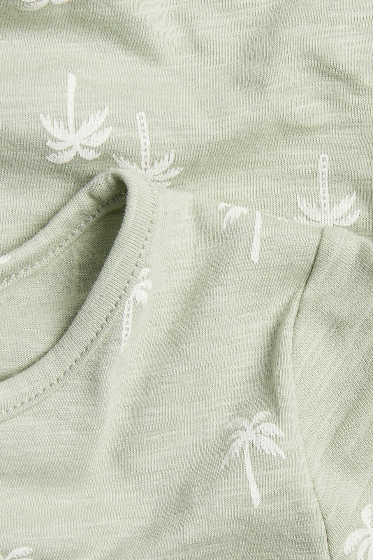 Zbliżenie na jasnozielony bawełniany pajacyk niemowlęcy z białym nadrukiem w palmy.