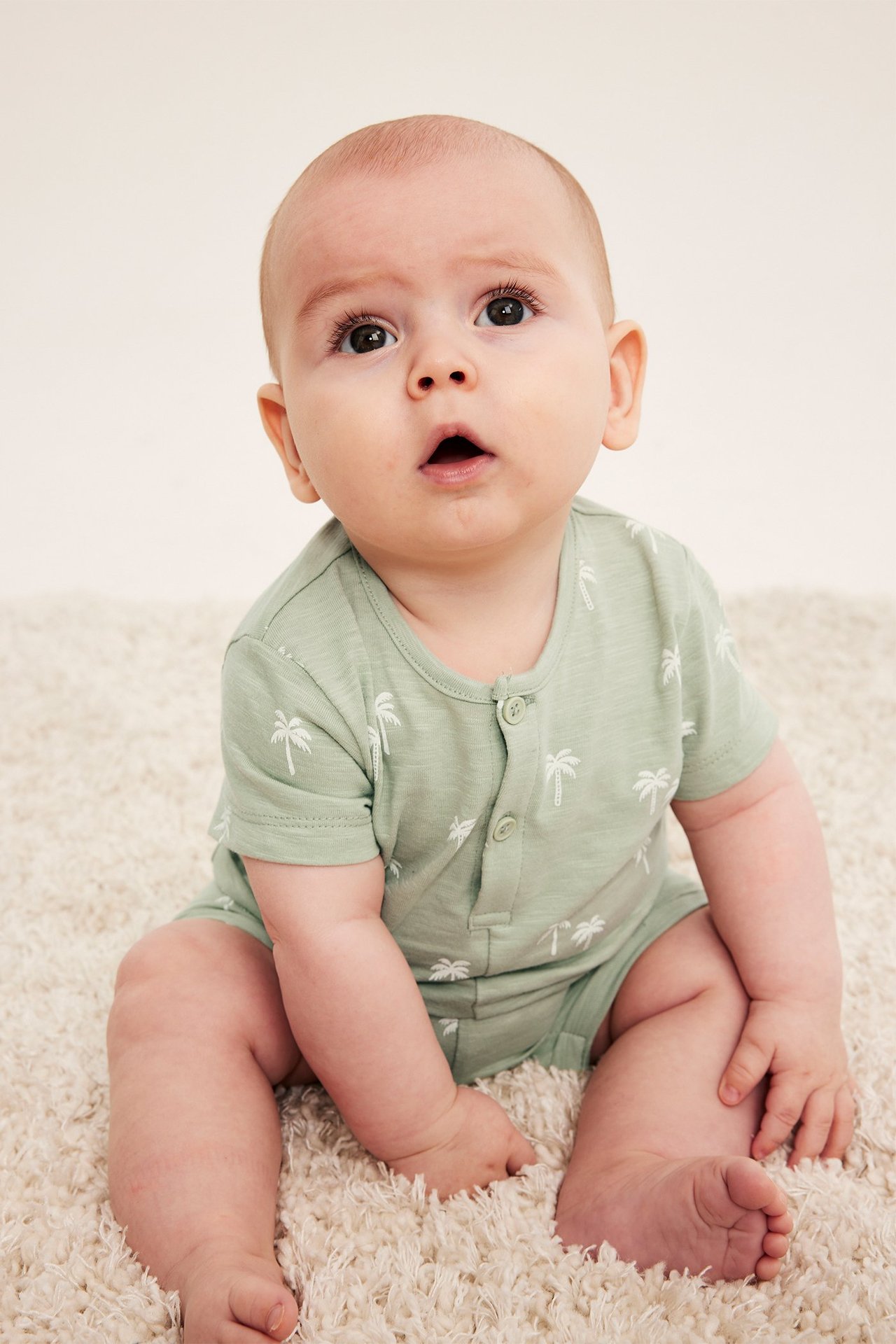 Widok z przodu dziecka w zielonym bawełnianym kombinezonie z krótkim rękawem, nadrukiem palm i guzikami.