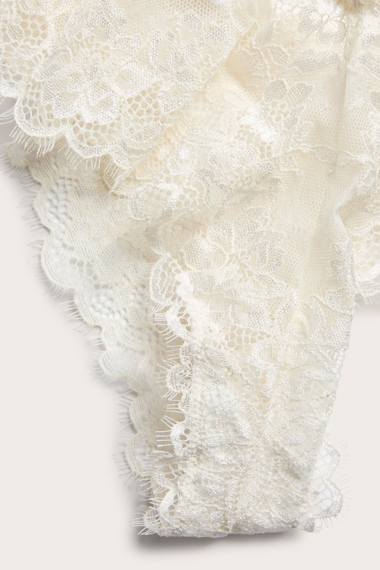 Zbliżenie na majtki typu cheeky w kolorze écru z koronki, z misternymi wzorami kwiatowymi wzdłuż falowanego brzegu.