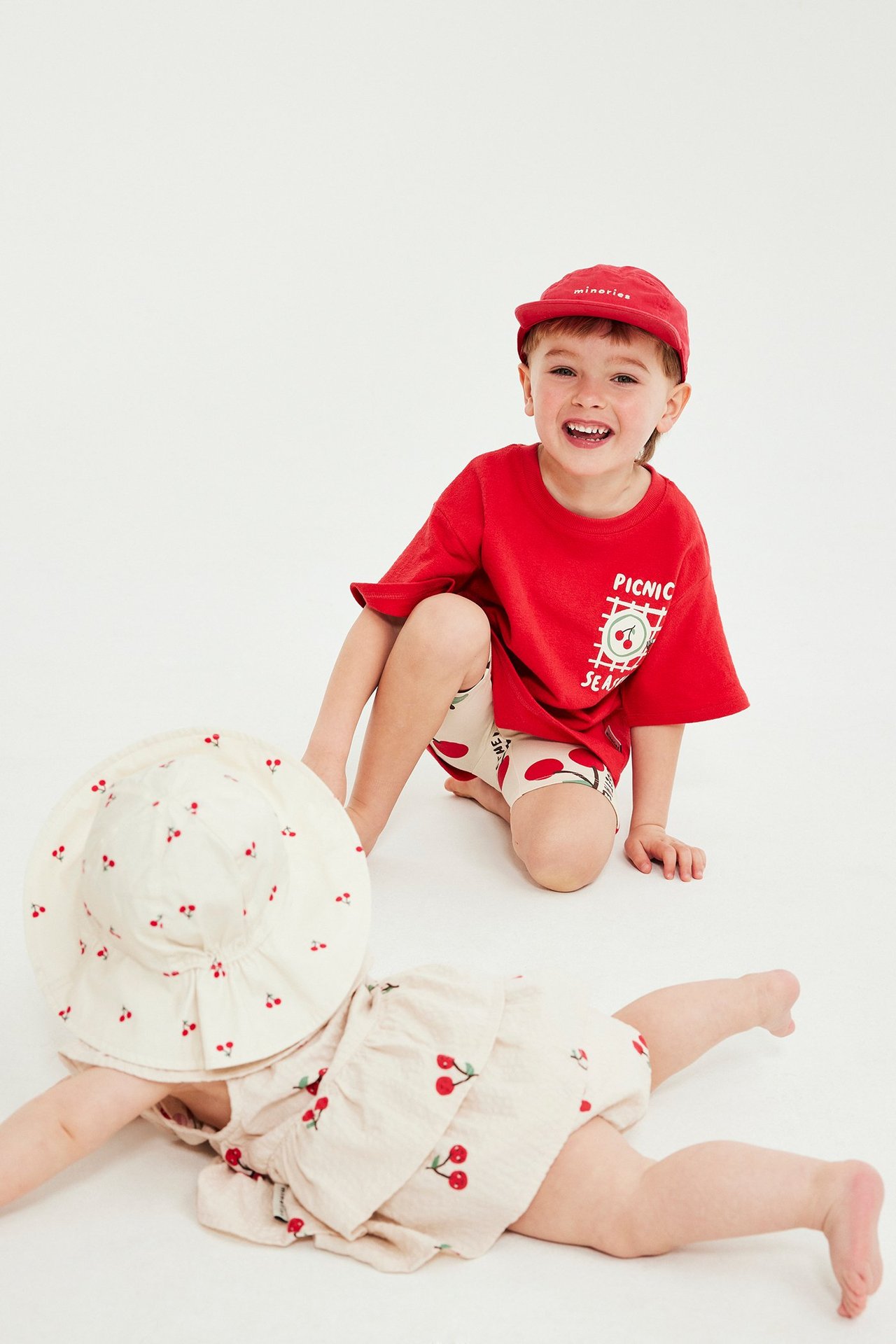 Barn i röd tryckt t-shirt, vita körsbärsshorts och röd keps. Baby i matchande körsbärsmönstrad sparkdräkt.