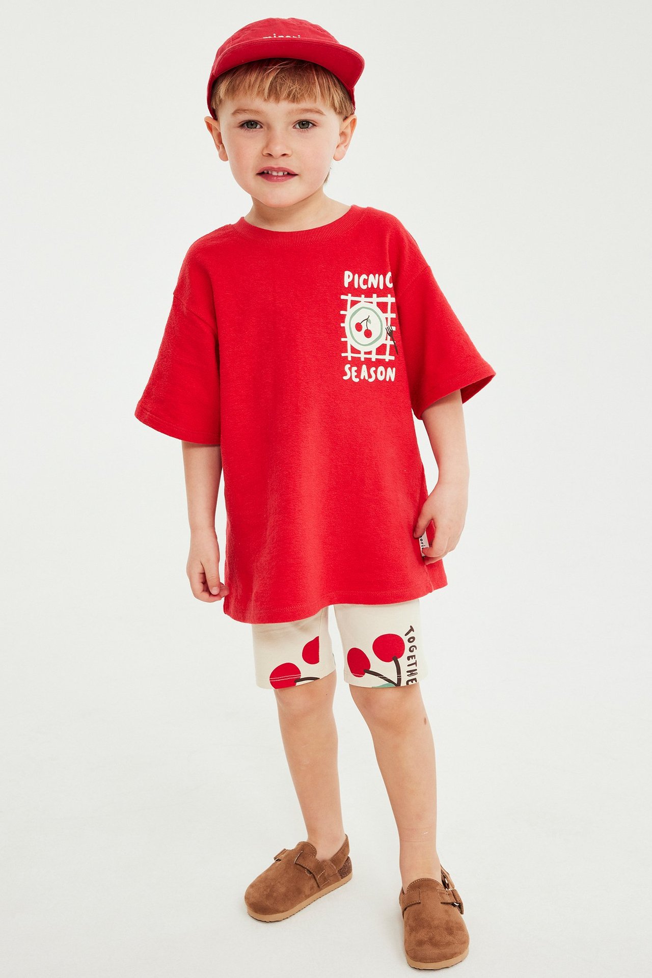 Dziecko w czerwonym t-shircie z wiśniami, kremowych szortach w wisienki, czerwonej czapce i brązowych butach.