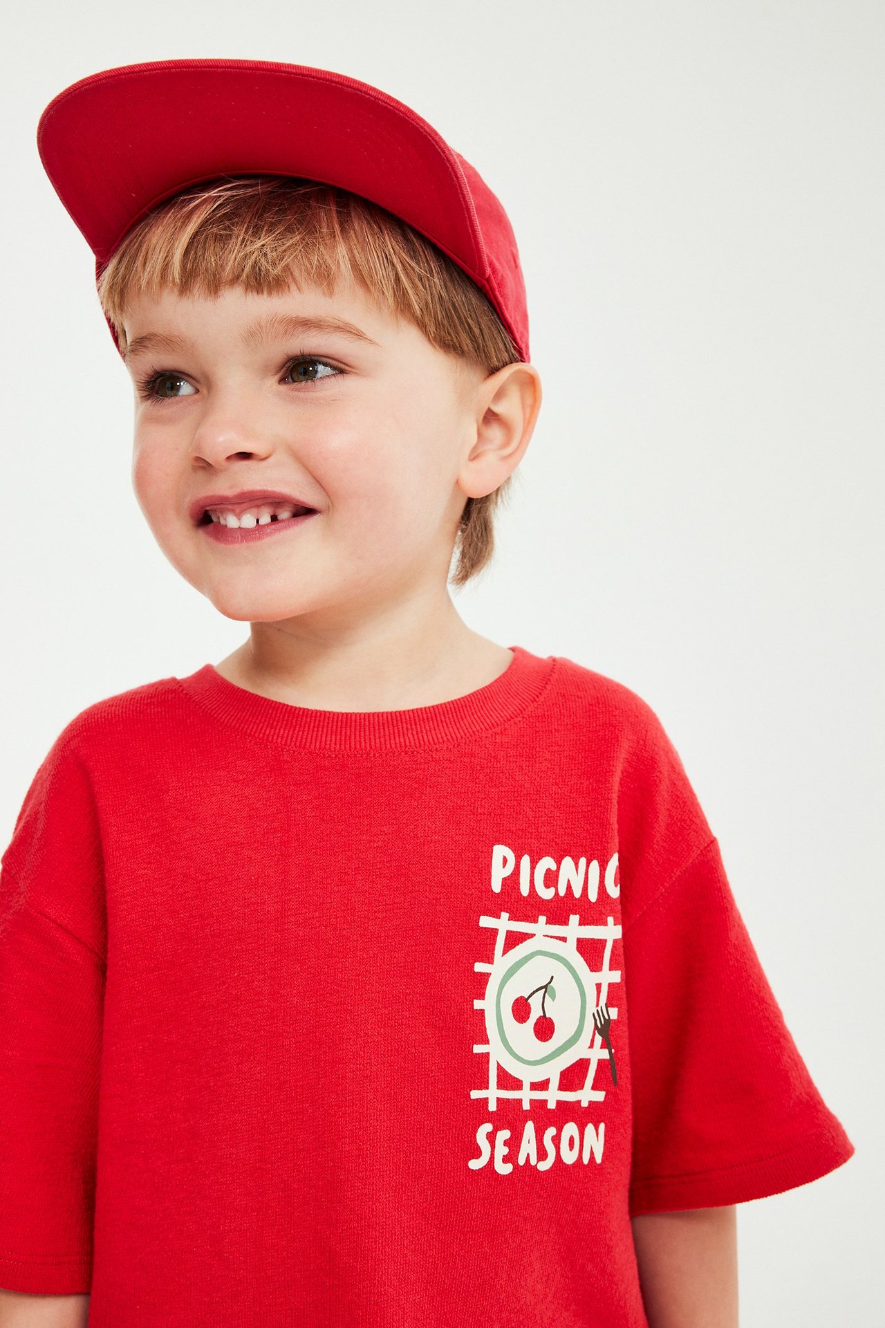 Widok z przodu dziecka w czerwonej koszulce z krótkim rękawem z nadrukiem wiśni i czerwonej czapce.