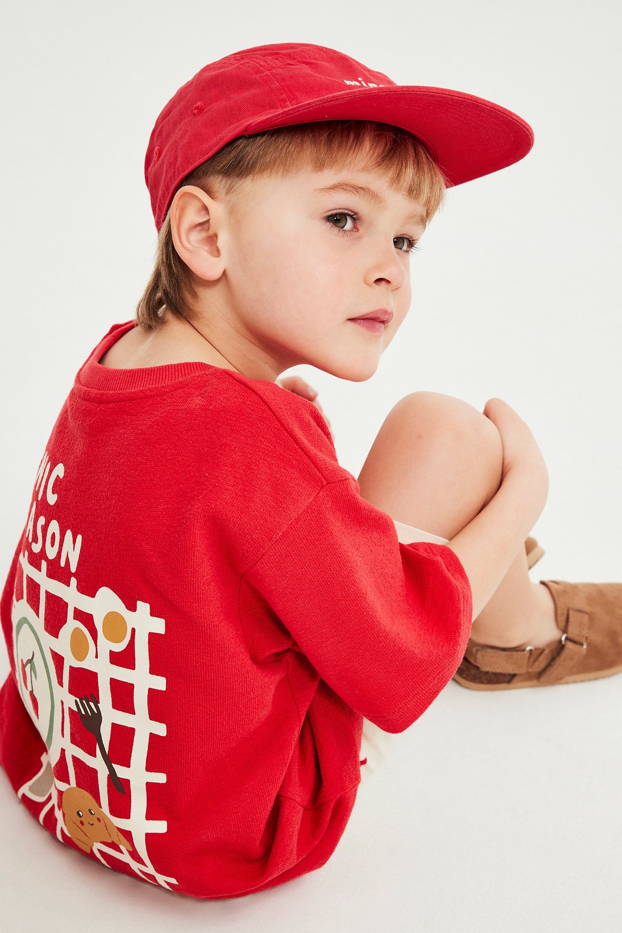 Sidovy av ett barn i en röd bomulls-t-shirt med matmönster, en röd keps och bruna sandaler.