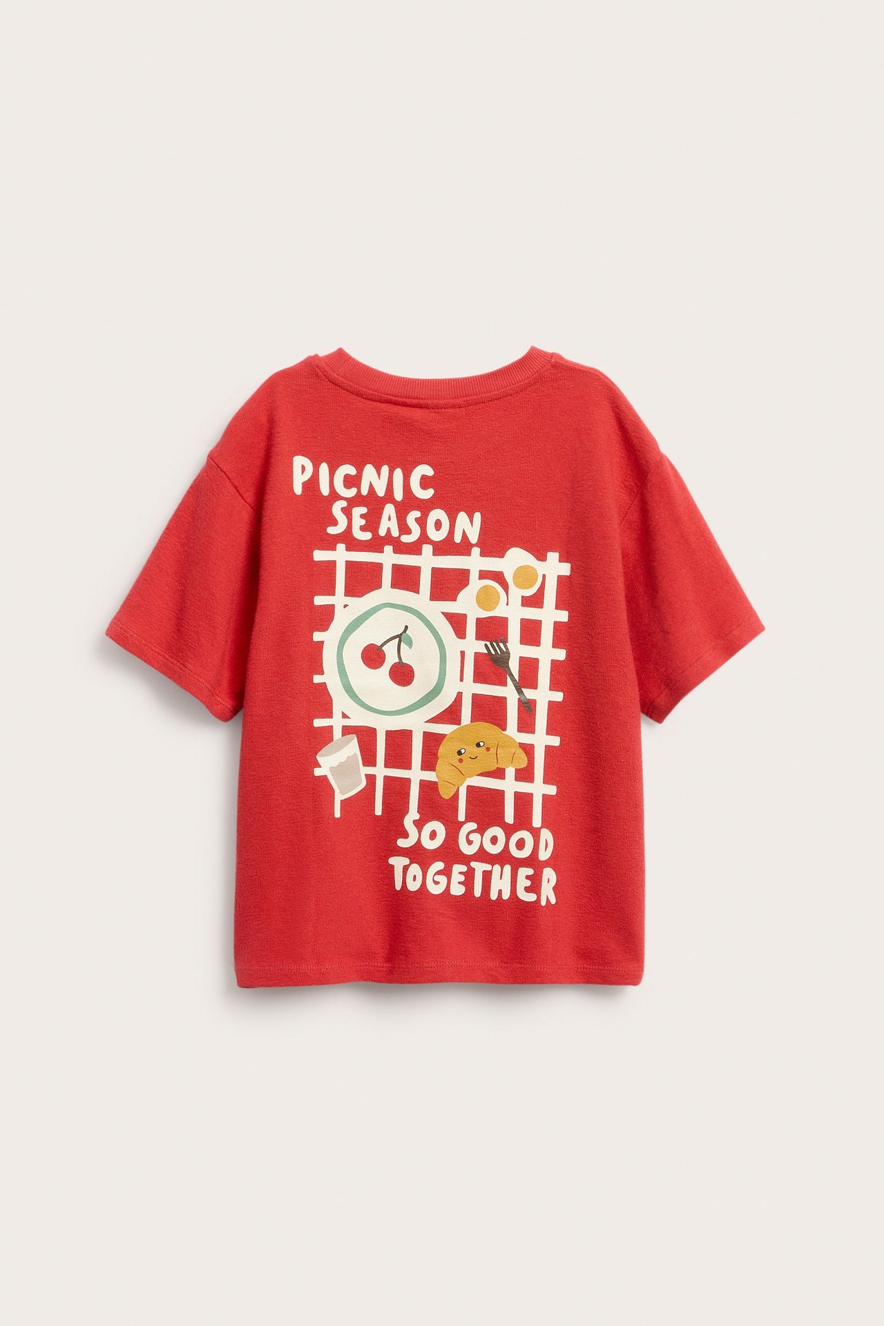 Frontvy av en röd bomullskortärmad t-shirt för barn med grafiskt tryck av picknickmat och text.