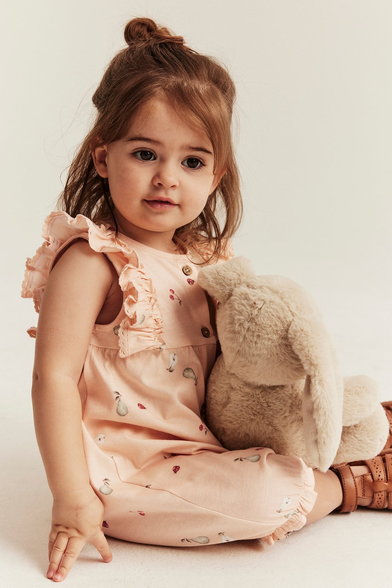 Vauva edestä, yllään vaaleanpunainen hedelmäkuvioitu hihaton romper, pitäen pörröistä beigeä pupua. Ruskeat sandaalit.