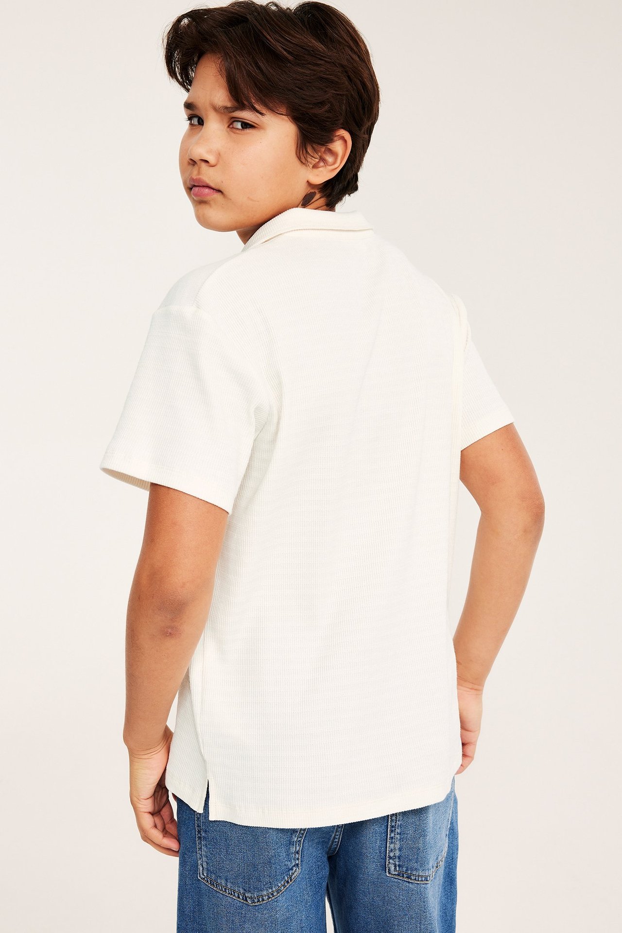 Lapsi takaapäin, yllään luonnonvalkoinen strukturoitu puuvillainen pikeepaita ja siniset farkut.