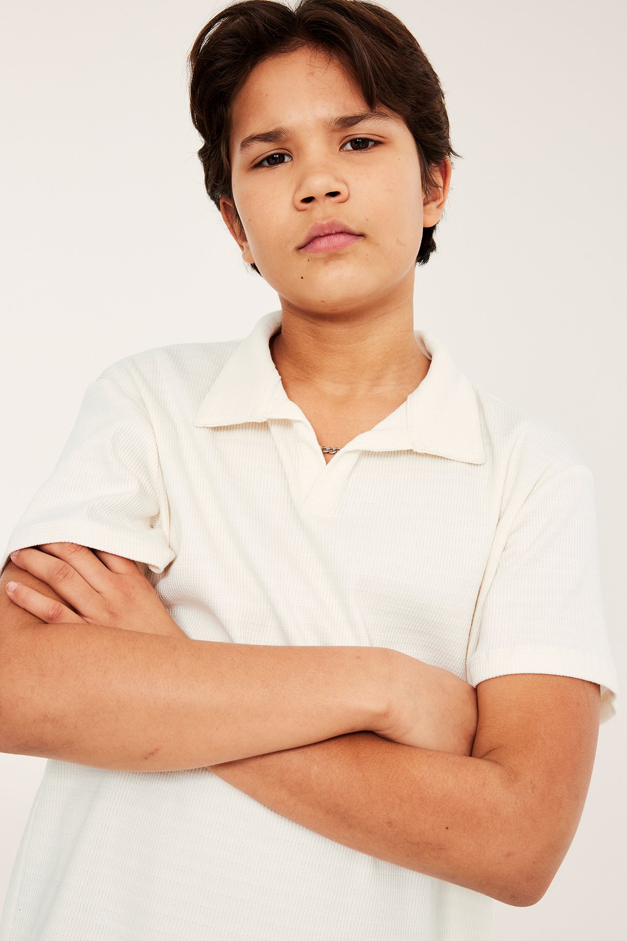 Edestä kuvattu lapsi yllään luonnonvalkoinen, rento, lyhythihainen teksturoitu poolopaita V-pääntiellä.