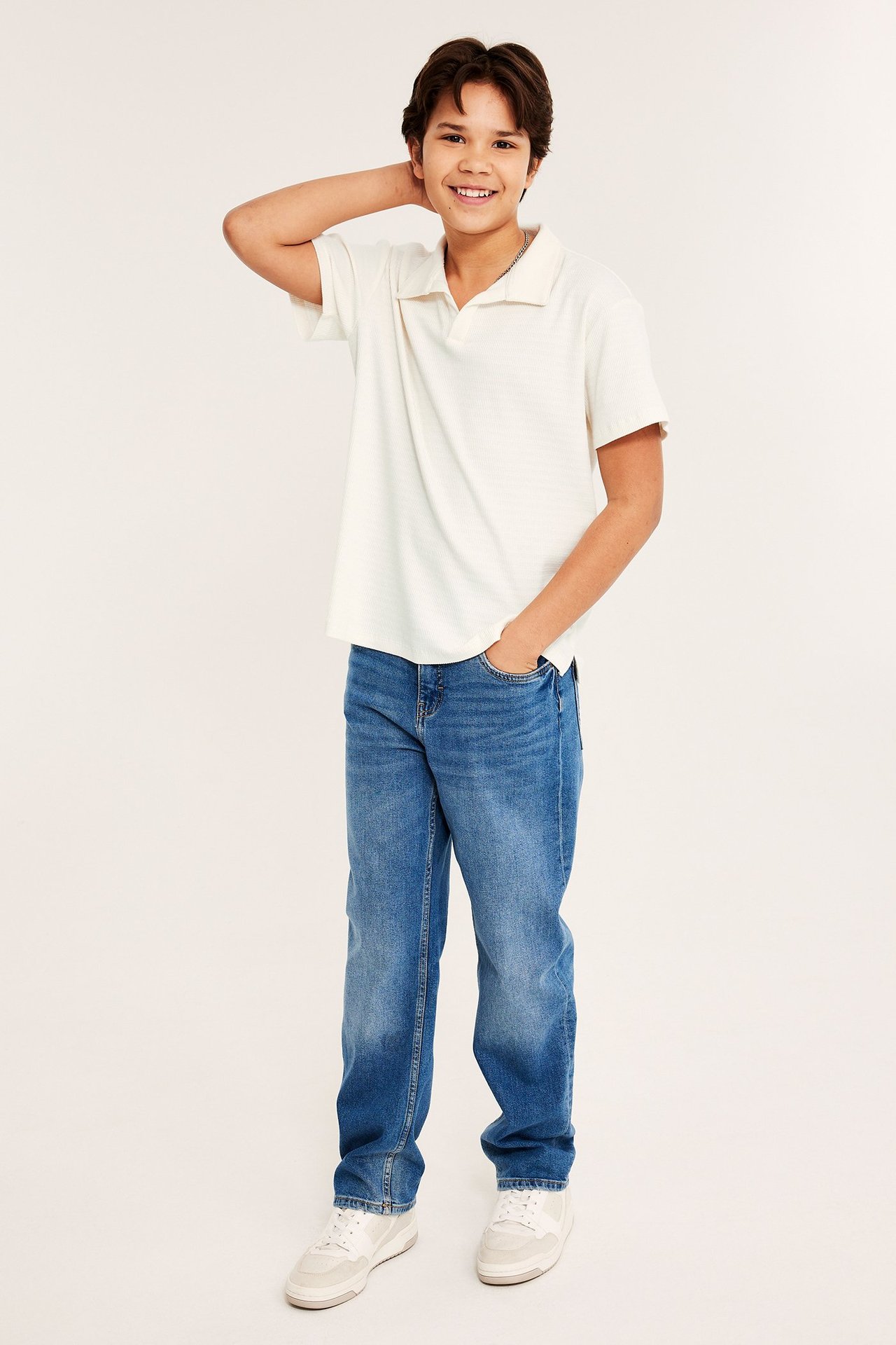 Edestä, hymyilevä lapsi yllään luonnonvalkoinen rento teksturoitu lyhythihainen pikeepaita, siniset farkut ja valkoiset lenkkarit.