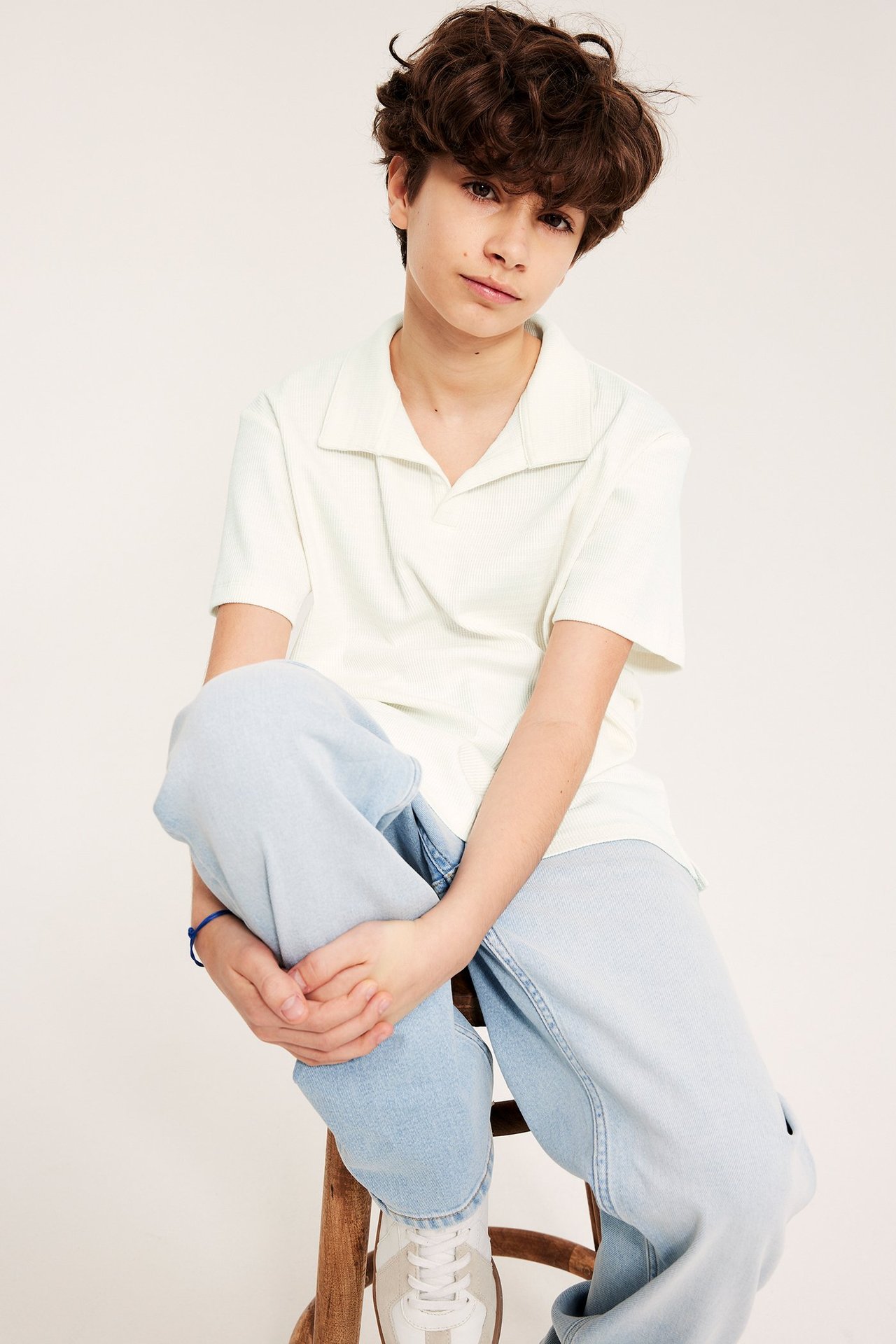 Edestäpäin: lapsi yllään offwhite lyhythihainen pikeepaita, vaaleansiniset farkut ja valkoiset tennarit.