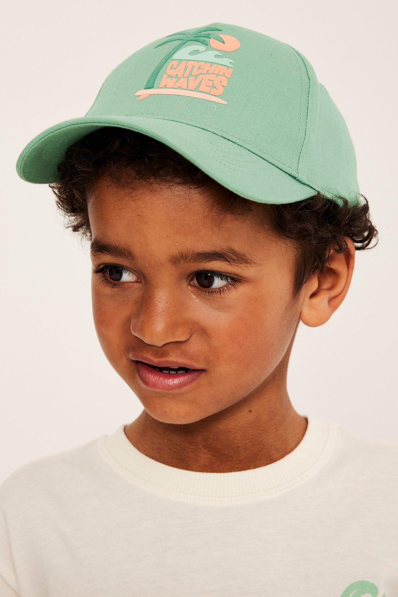 Lähikuva lapsesta, jolla on vaaleanvihreä puuvillatvillilippalakki, jossa on "Catchin' Waves" kohopainatus.
