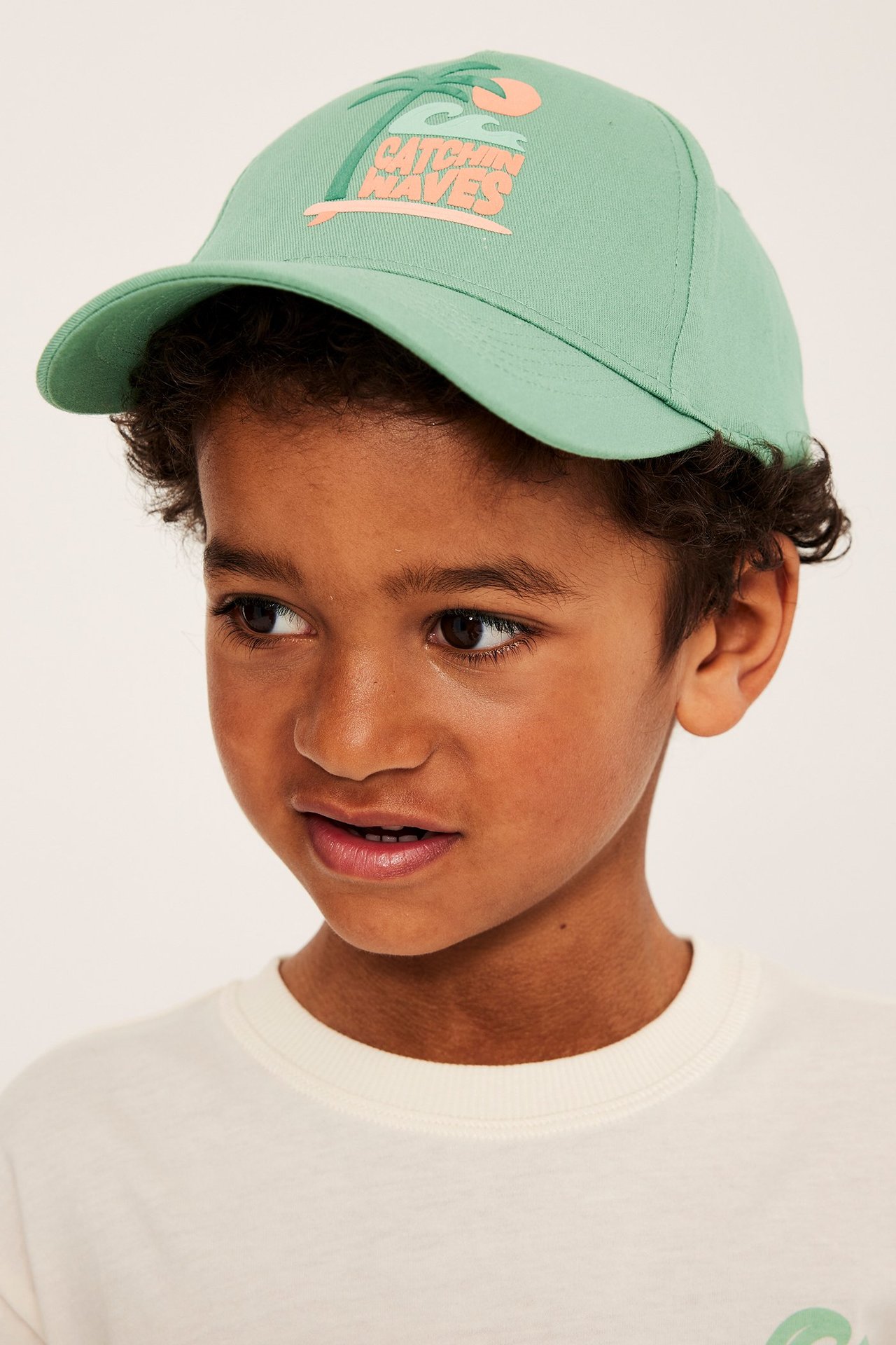 Lähikuva lapsesta, jolla on vaaleanvihreä puuvillatvillilippalakki, jossa on "Catchin' Waves" kohopainatus.