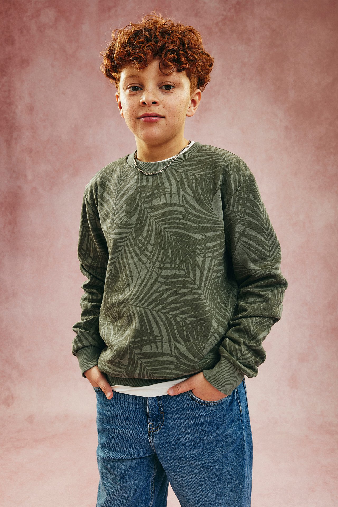 Edestäpäin, lapsi yllään vihreä kuviollinen froteepaita palmulehdillä, siniset farkut ja hopeakaulakoru.