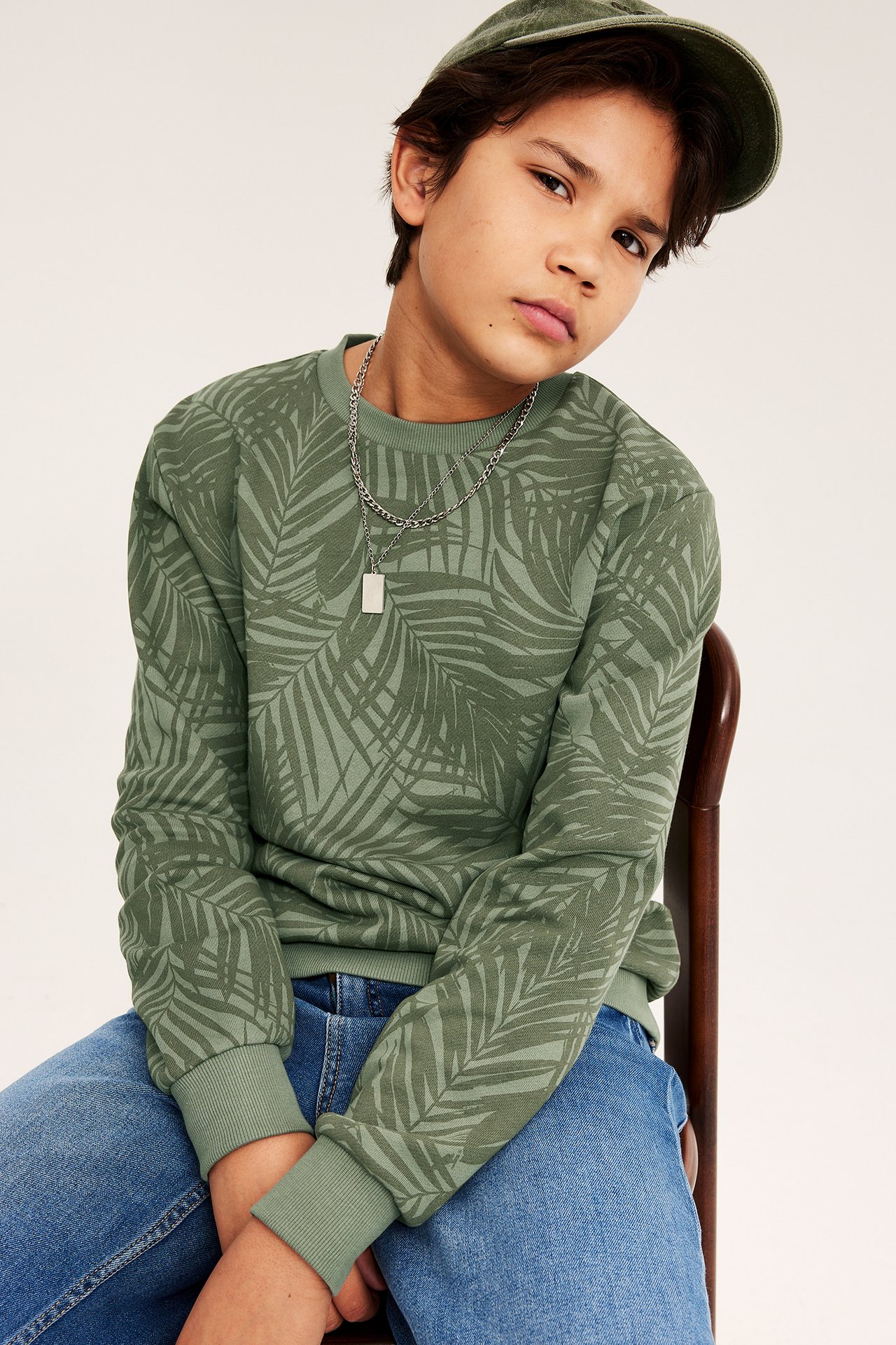 Edestäpäin, lapsi vihreässä kuviollisessa puuvillafleecepusero, sinisissä farkuissa, vihreä lippis, hopeaketju.