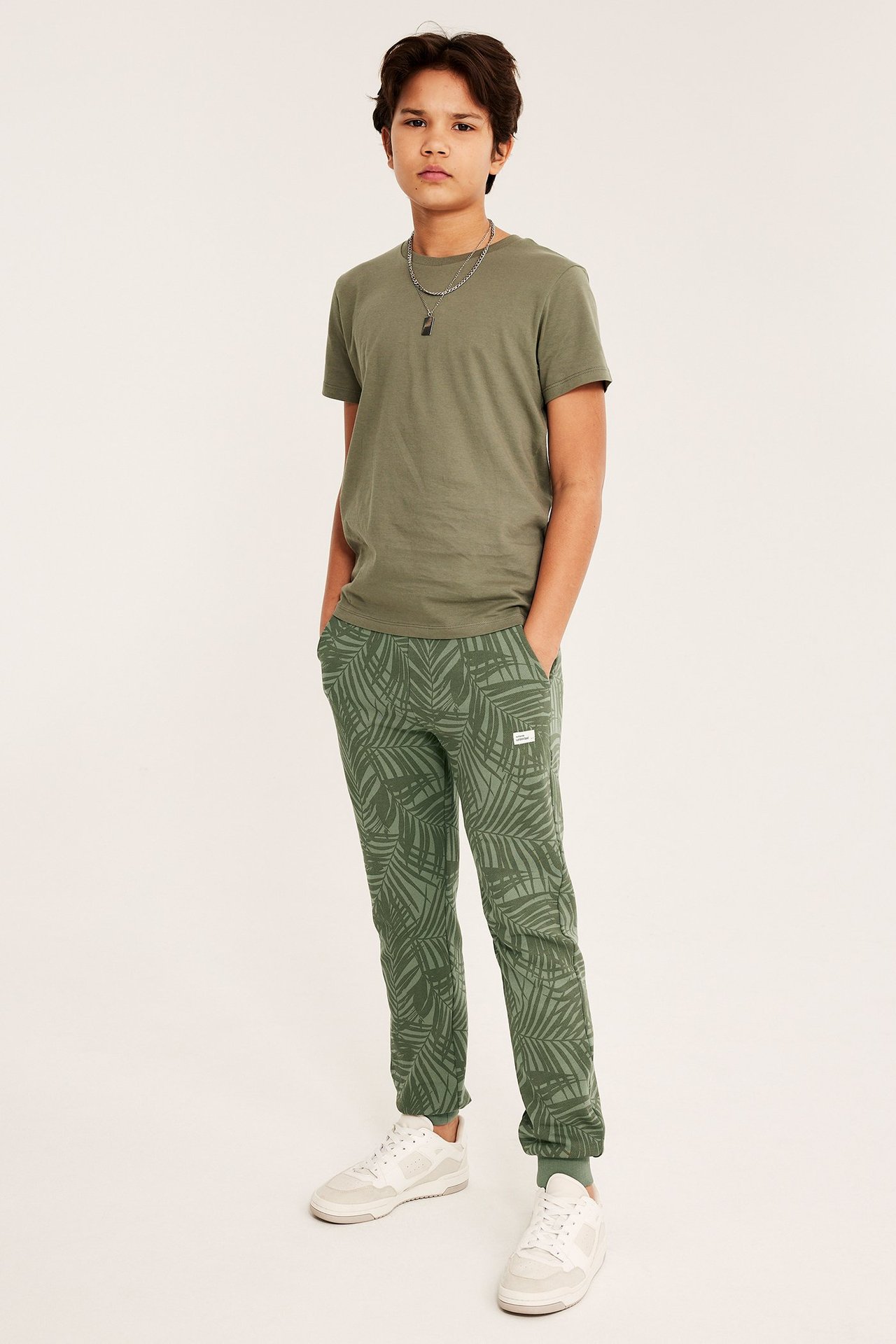 Chłopiec w zielonych wzorzystych spodniach dresowych, oliwkowej koszulce i białych trampkach.