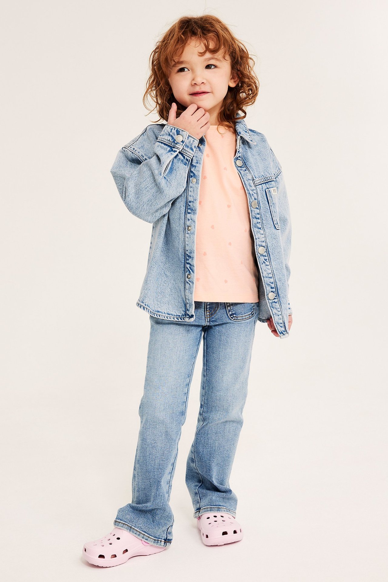 Framifrån: Ett barn i ljusorange hjärtmönstrad bomullstopp, ljusblå jeansjacka, jeans och rosa träskor.