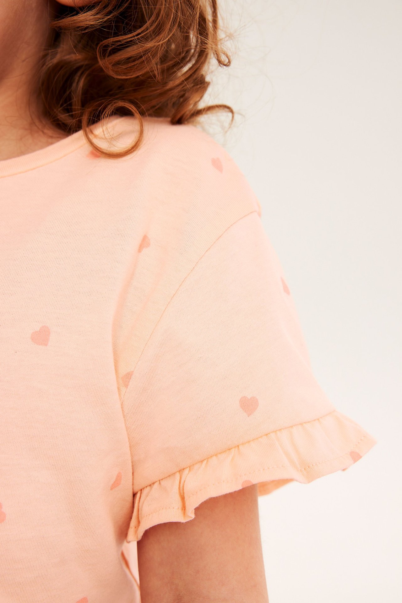 Lähikuva lapsesta yllään vaaleanoranssi sydänkuvioinen puuvillainen t-paita röyhelöhihalla.
