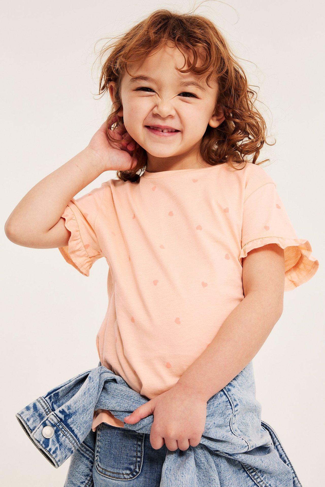 Frontbild: Barn i ljusorange hjärtmönstrad bomullstopp med volangärmar, denimjacka knuten i midjan.