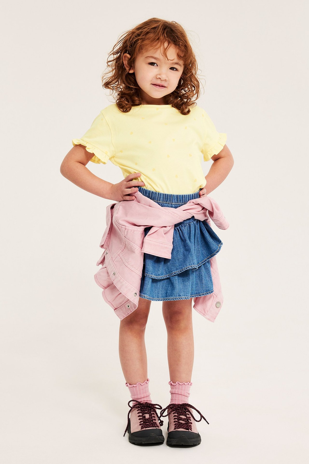 Frontvy: Barn bär ljusgul hjärtmönstrad bomullstopp med volangärm, blå jeanskjol, rosa jacka och skor.