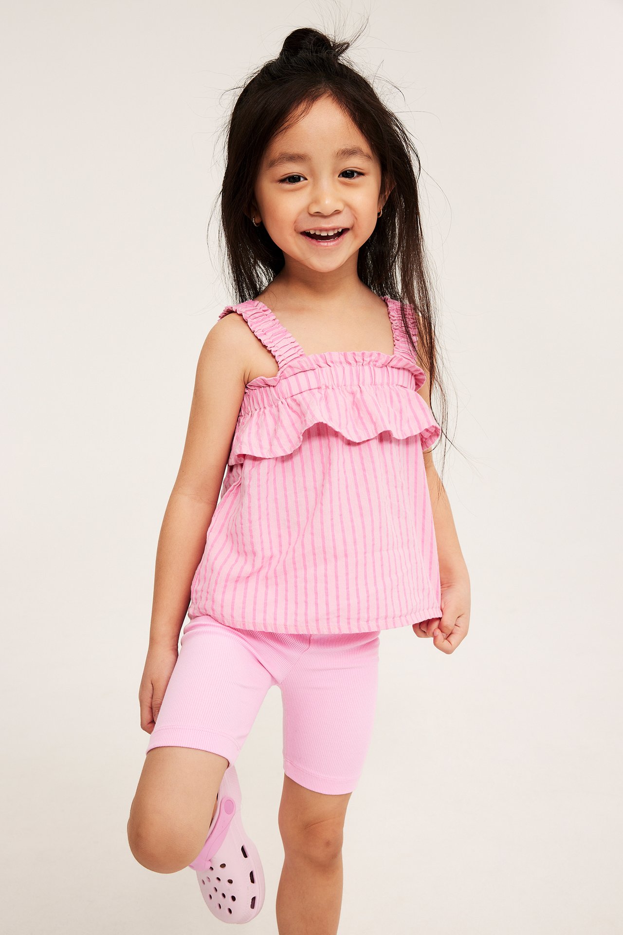 Forfra: barn i rosa ribbestrikket sykkelshorts, en rosa stripete volangtopp og rosa tresko.
