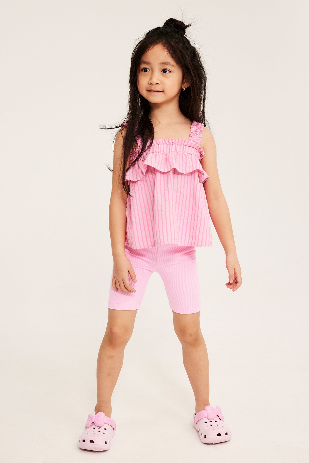 Frontvisning. Barn i lyserosa ribbestrikket sykkelshorts, rosa stripet volangtopp og rosa sandaler.