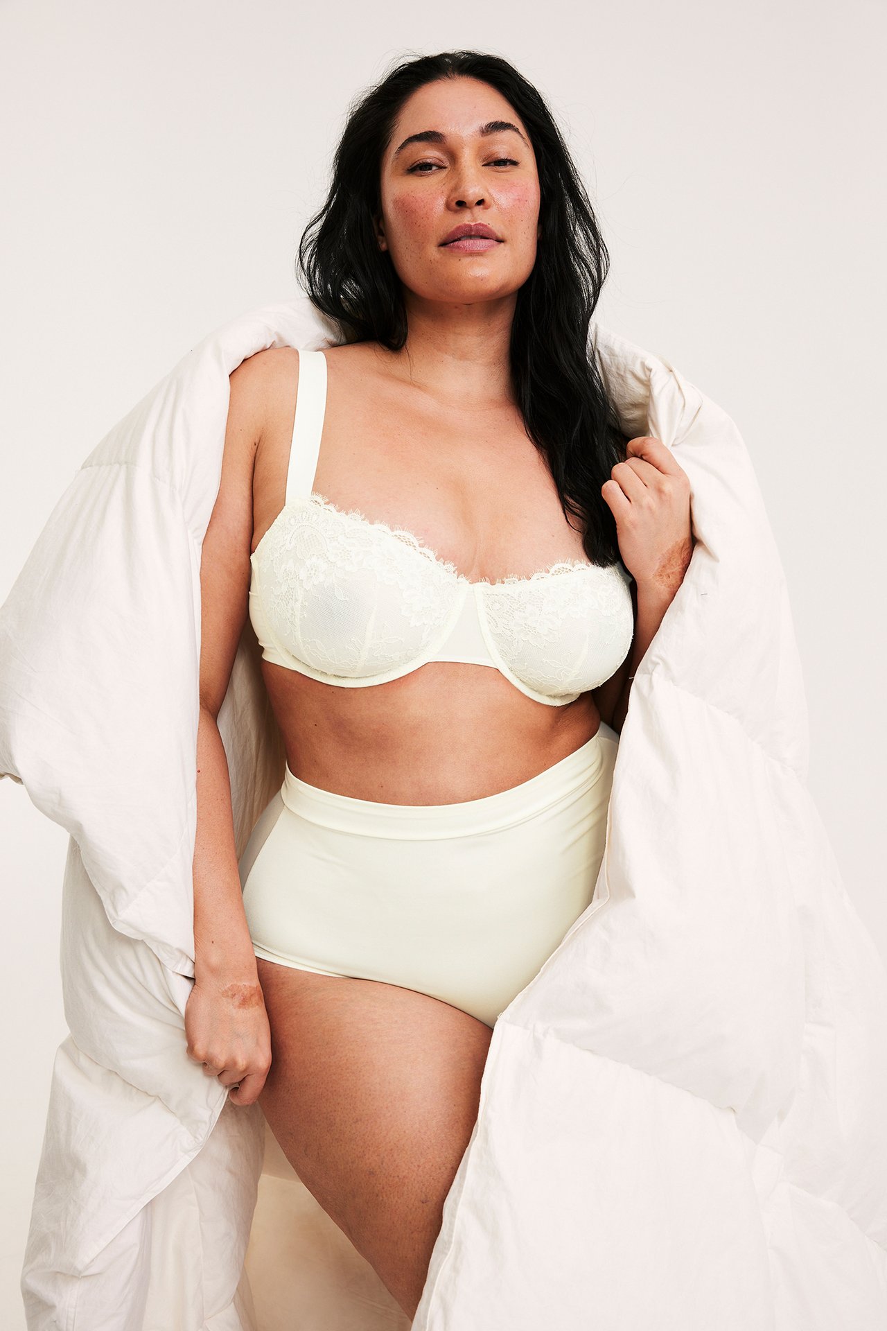 Etunäkymä naisesta, jolla on luonnonvalkoiset korkeavyötäröiset muotoilevat alushousut ja yhteensopivat pitsirintaliivit.