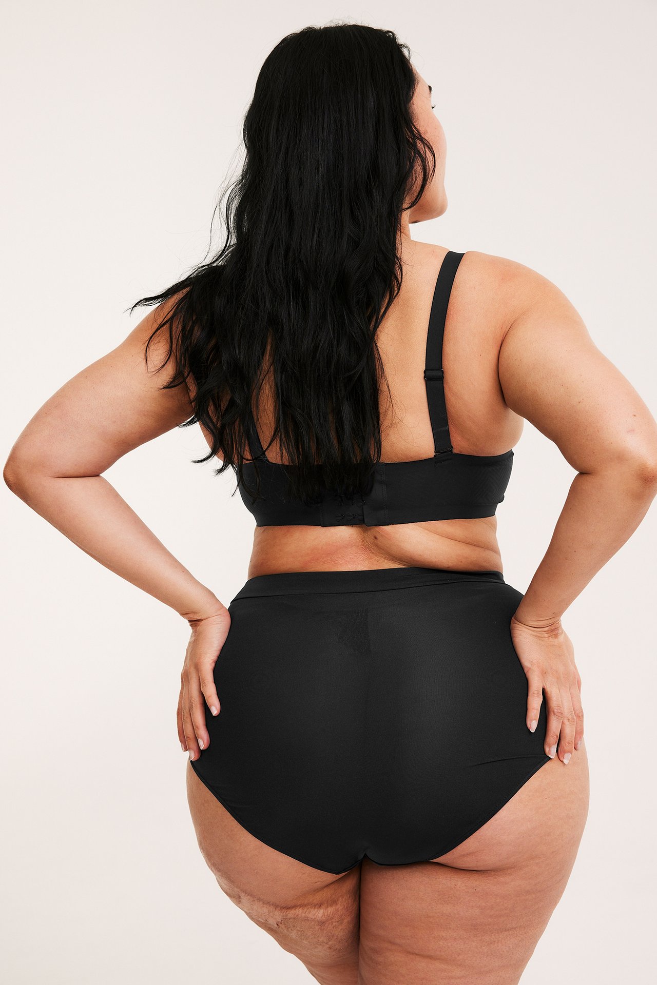 Takakuva naisesta yllään mustat korkeavyötäröiset muotoilevat alushousut ja mustat rintaliivit.