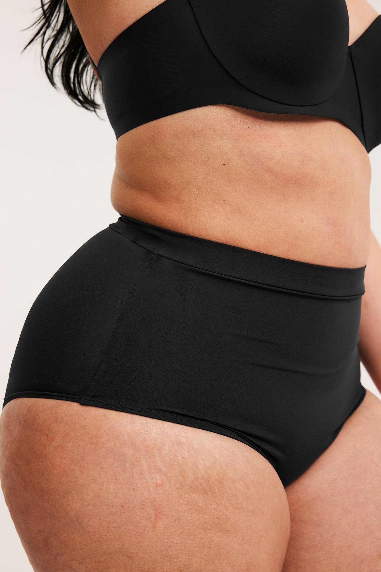 Sivukuva naisesta mustissa korkeavyötäröisissä muotoilevissa alushousuissa ja mustissa olkaimettomissa rintaliiveissä.