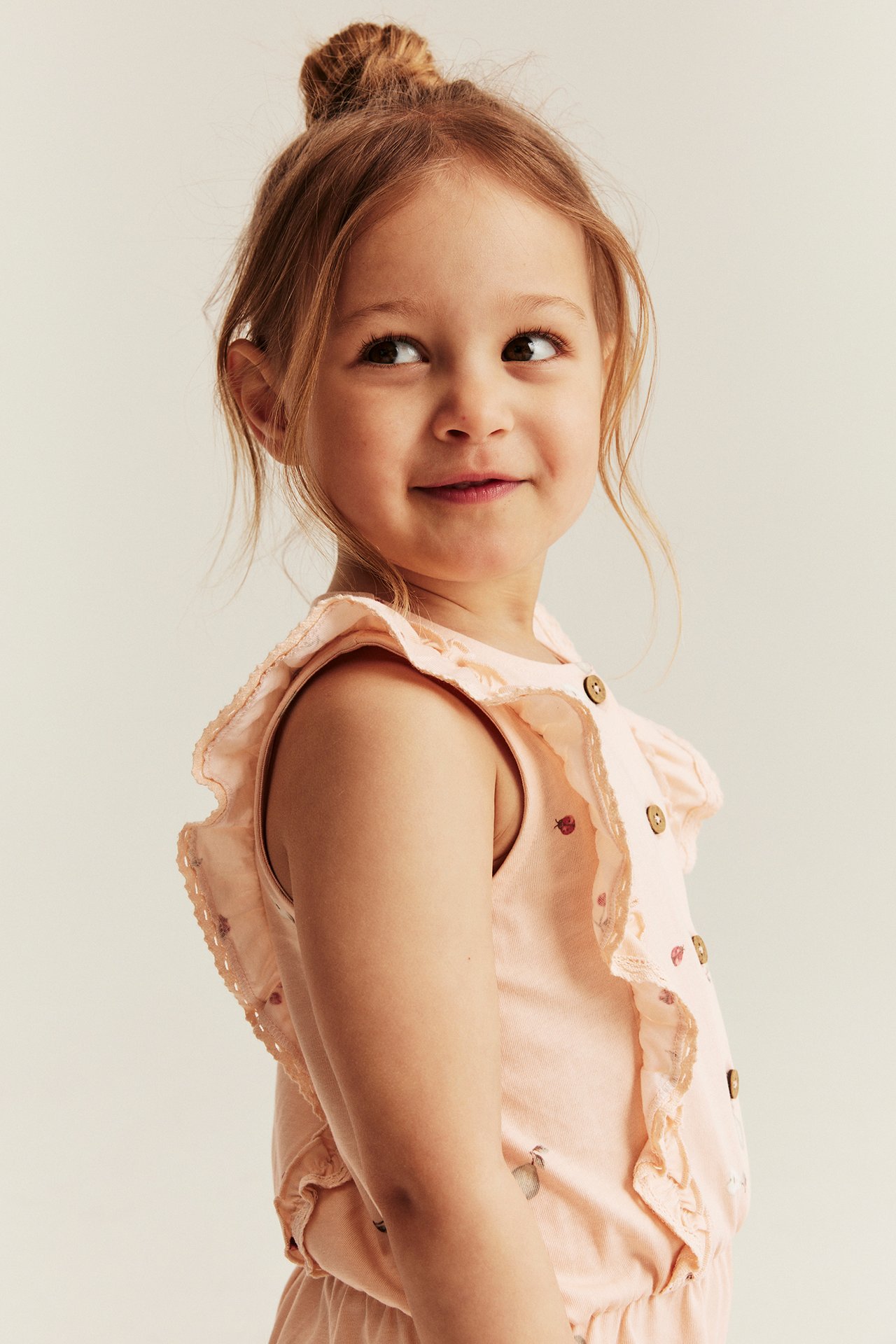 Lähikuva: Lapsi yllään vaaleanoranssi kuvioitu puuvillahaalari röyhelöillä, pitsillä ja napeilla.