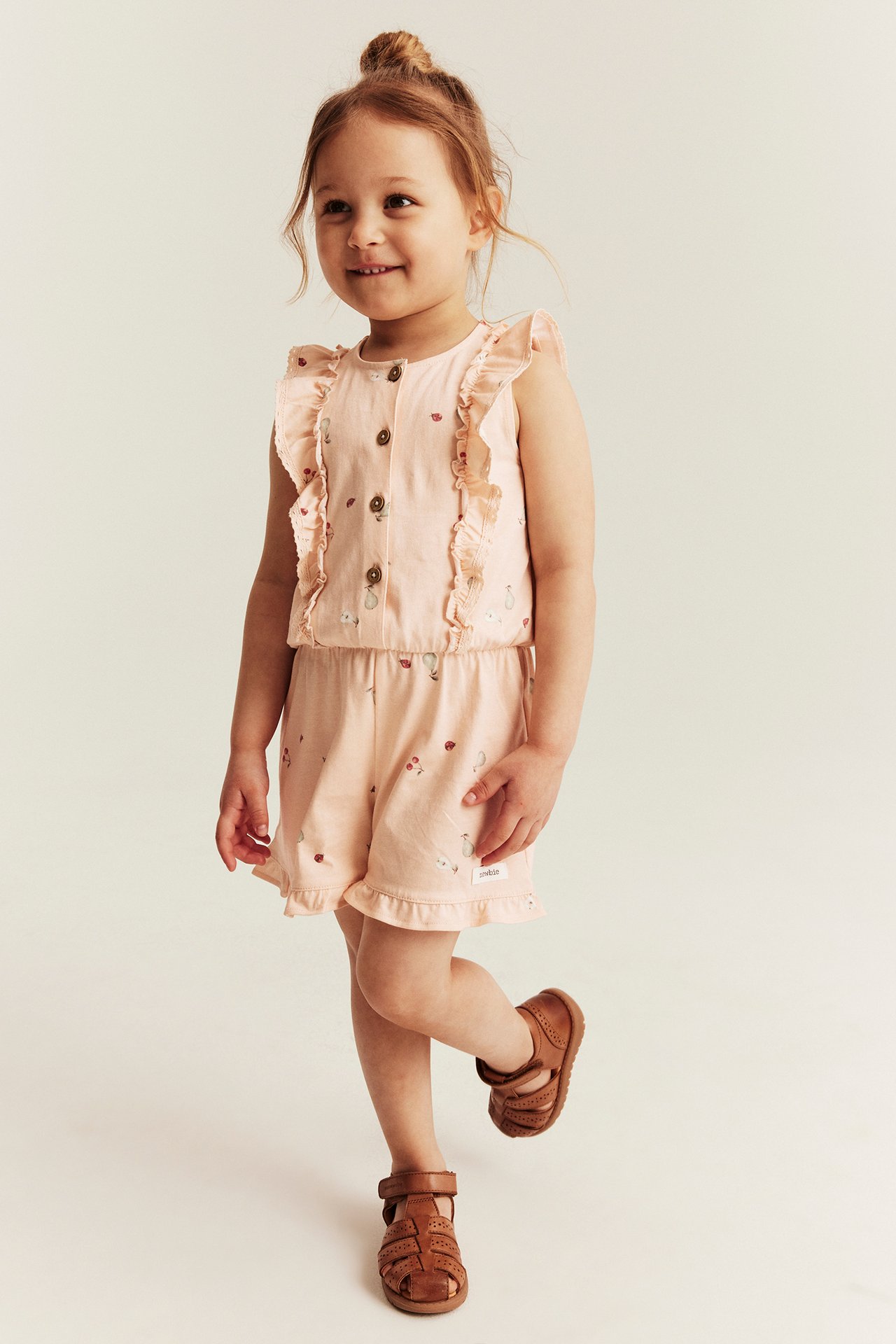 Edestä: Lapsi vaaleassa persikanvärisessä kuviollisessa jerseyhaalarissa, jossa röyhelöitä ja napit, ruskeat sandaalit jalassa.