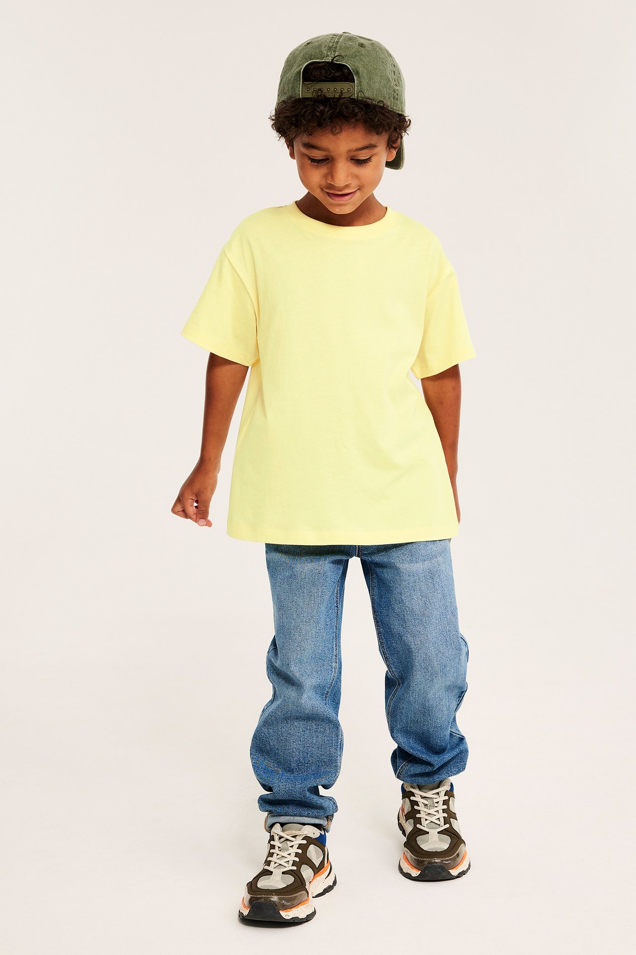 Framifrån: ett barn i gul bomulls-T-shirt, blå jeans, olivgrön keps och flerfärgade sneakers.