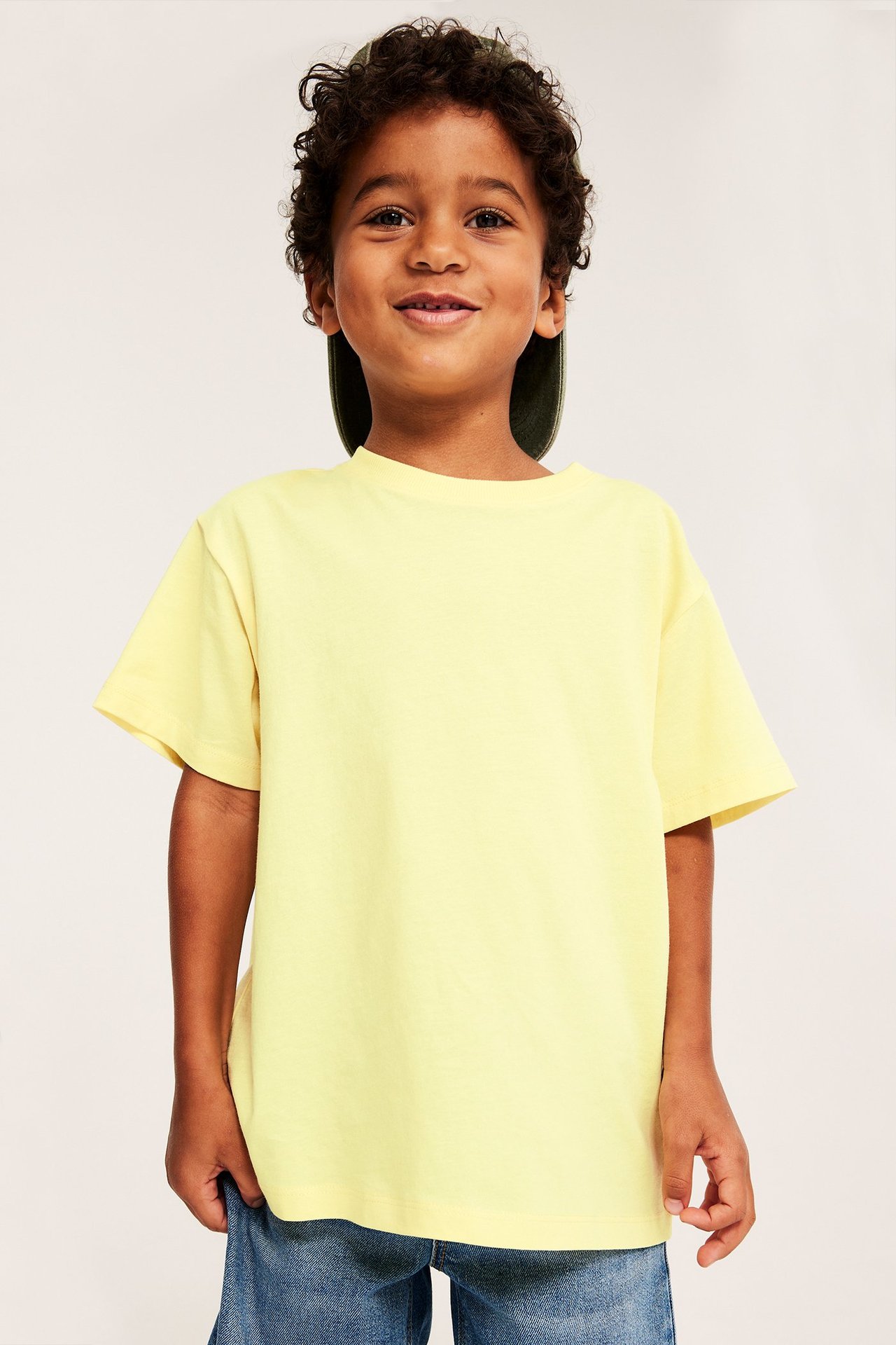 Framifrån, ett leende barn i en gul kortärmad bomulls-t-shirt, blå jeans och en grön keps.