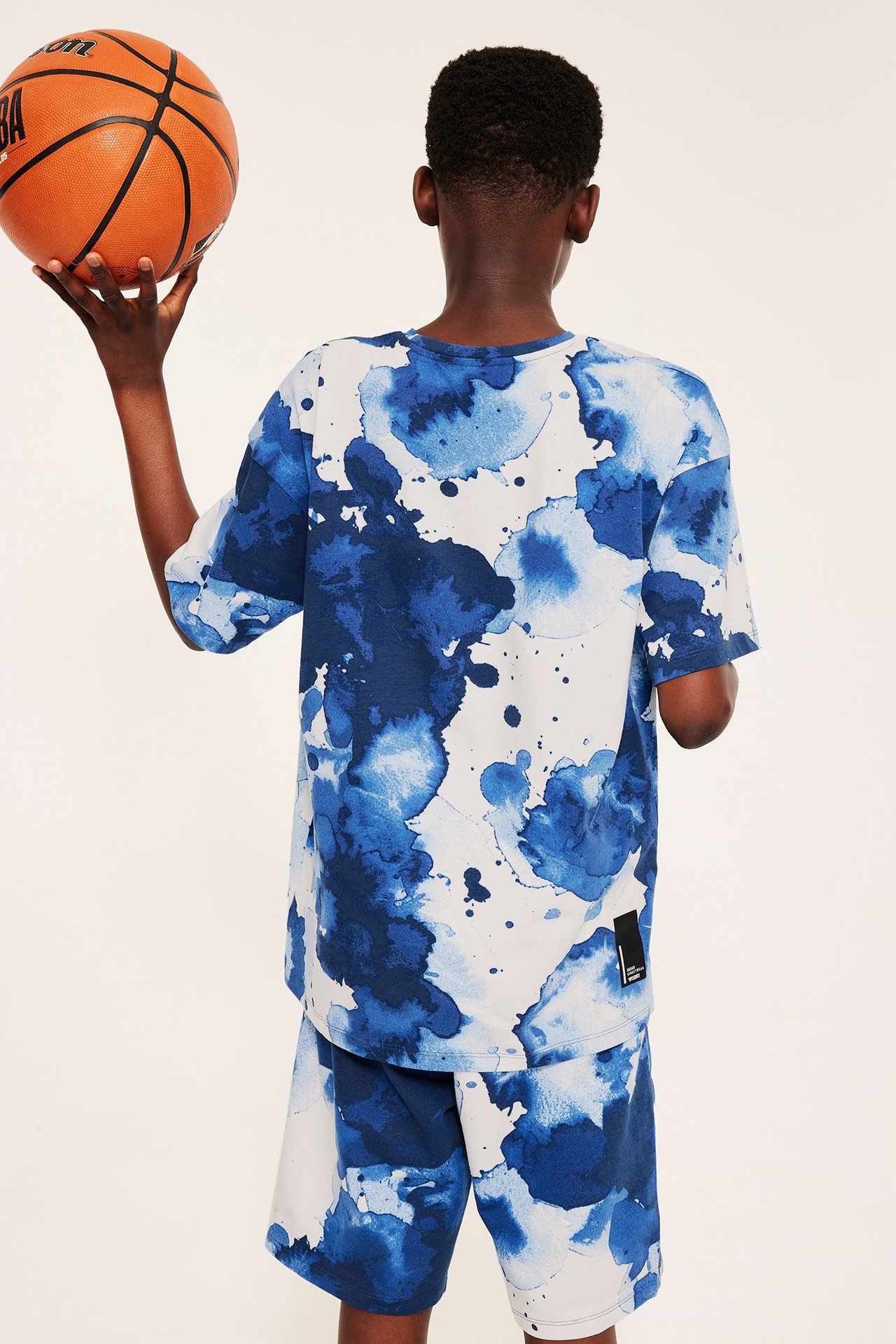 Lapsi takaa, ylisuuri luonnonvalkoinen puuvillainen t-paita sinisellä abstraktilla kuviolla ja shortsit, kädessään koripallo.
