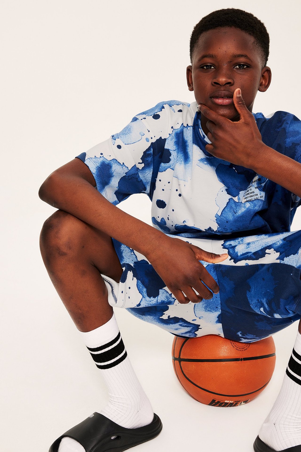 Framifrån, kid i oversized offwhite och blå abstrakt bomullst-shirt, matchande shorts, vita strumpor, svarta tofflor.