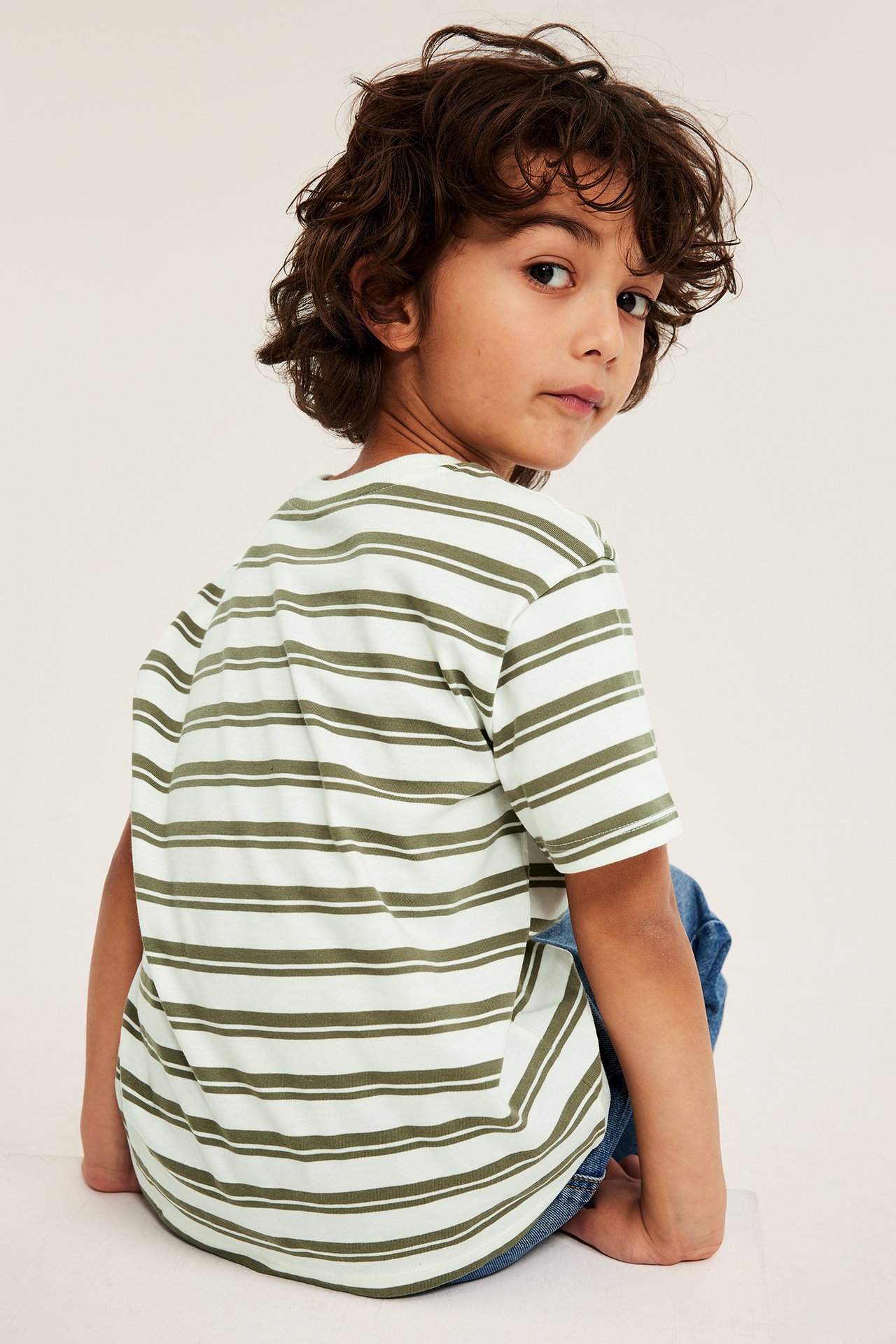 Ryggside-utsikt av et barn i en off-white og olivengrønn stripete oversized t-skjorte og blå jeans.