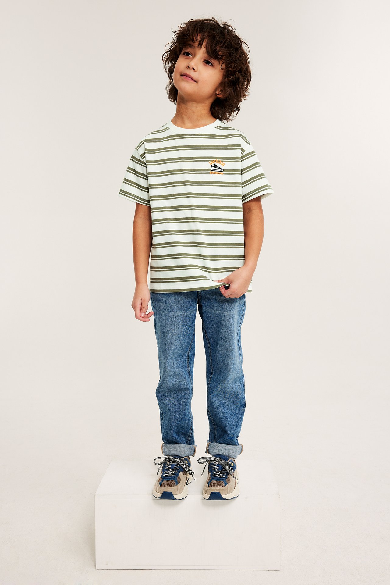 Front: Barn i offwhite, grønnstripet oversized bomulls-t-skjorte, blå jeans og joggesko.