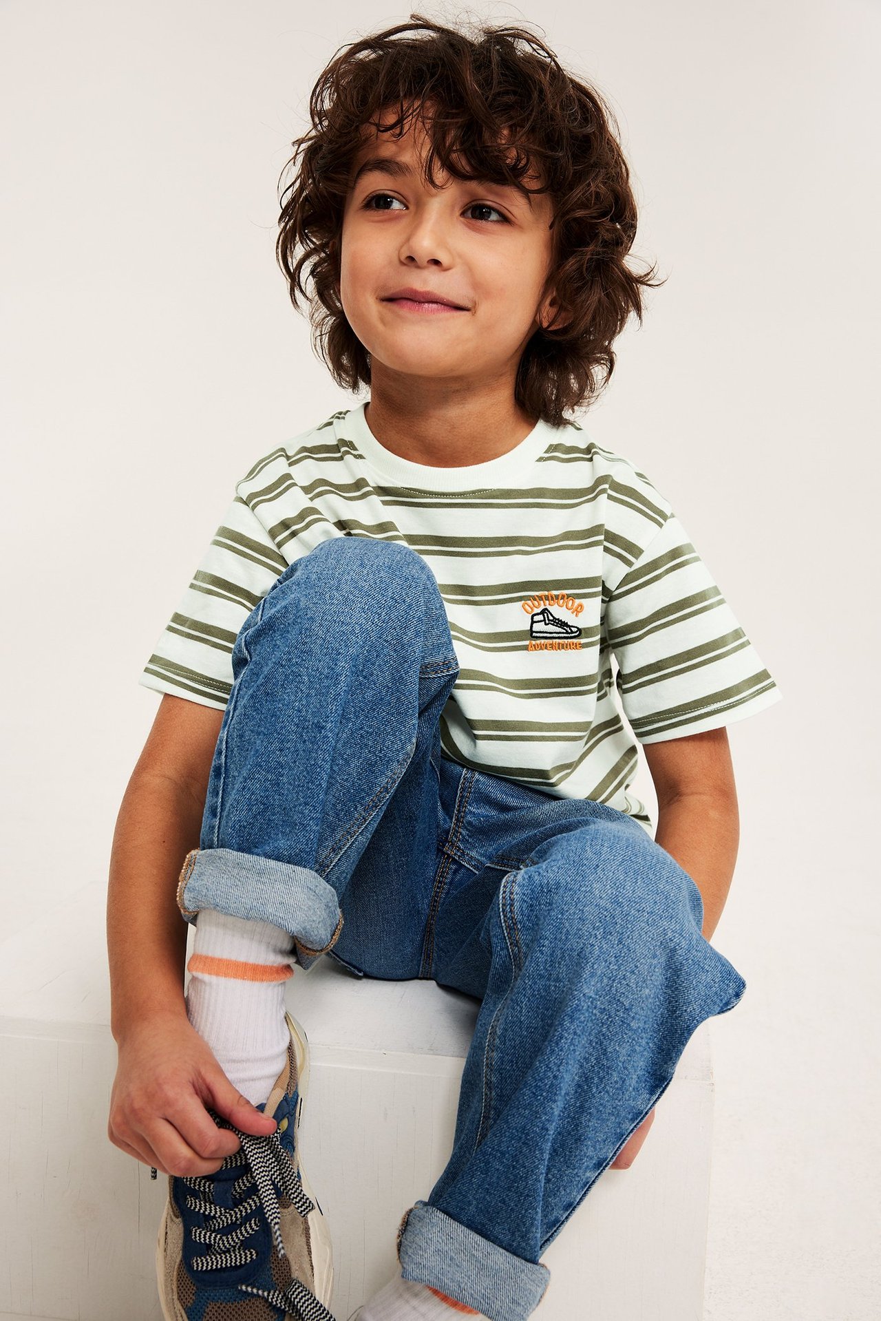 Framifrån: Barn i offwhite, olivrandig oversized t-shirt med broderi, blå jeans, vita strumpor och sneakers.