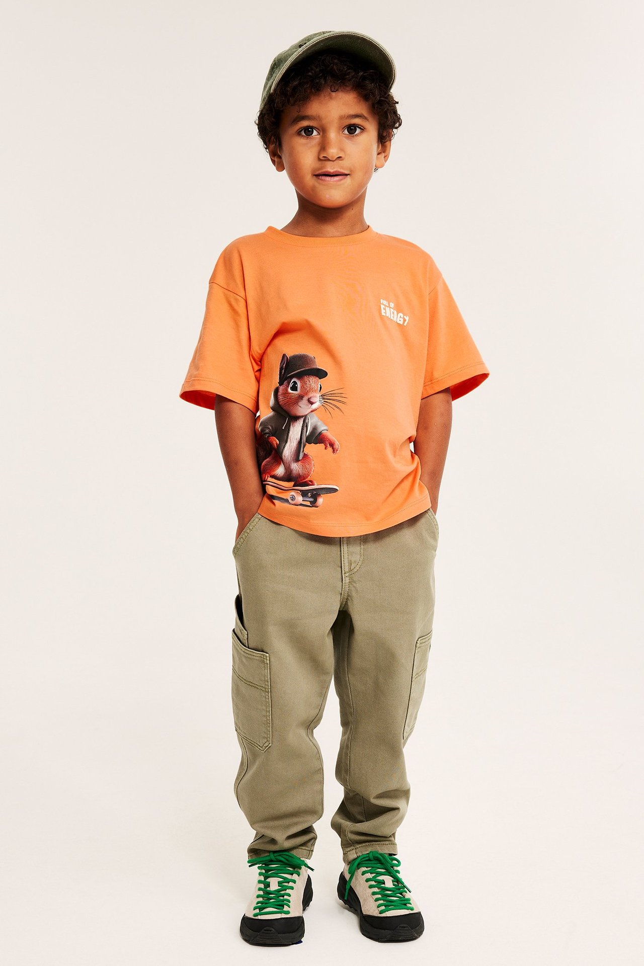 Lapsen oranssi puuvilla-T-paita oravaprintillä, oliivinvihreä lippalakki, khakihousut ja tennarit.