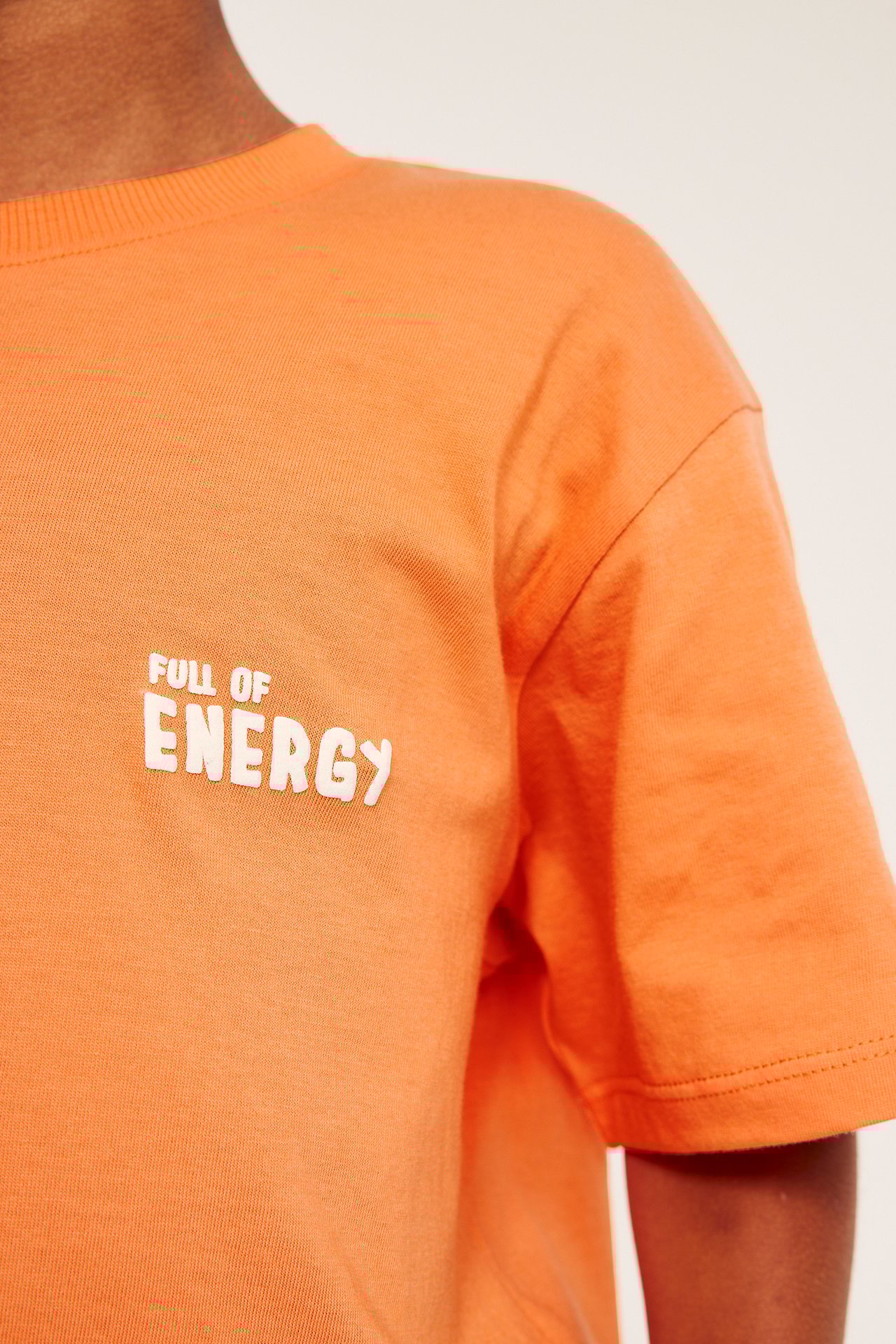 Närbild framifrån på ett barn i orange bomulls-t-shirt med vitt 'FULL OF ENERGY'-texttryck.