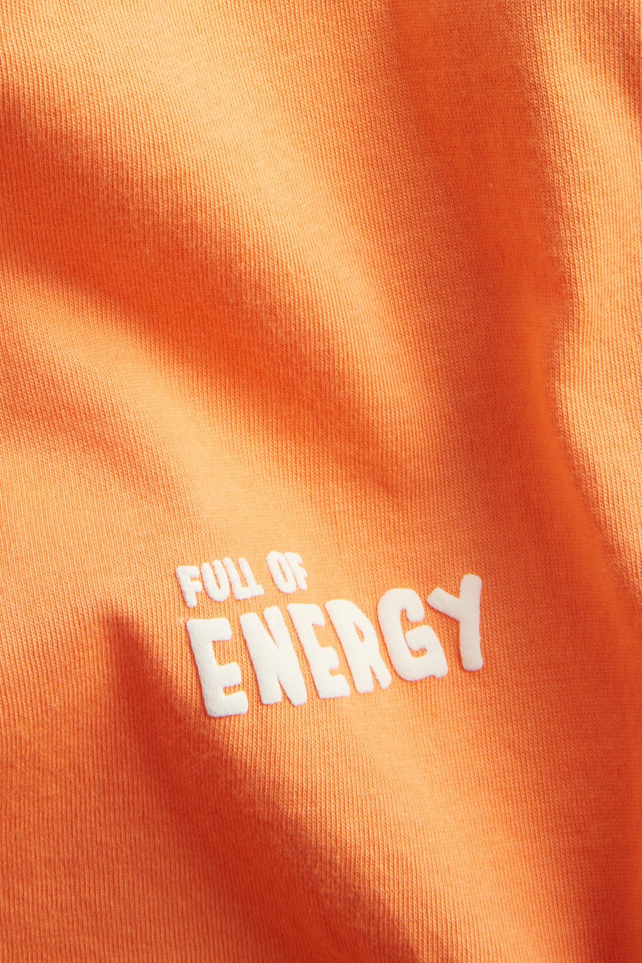 Lähikuva oranssista puuvilla T-paidasta, jossa valkoinen "FULL OF ENERGY" -tekstipainatus.