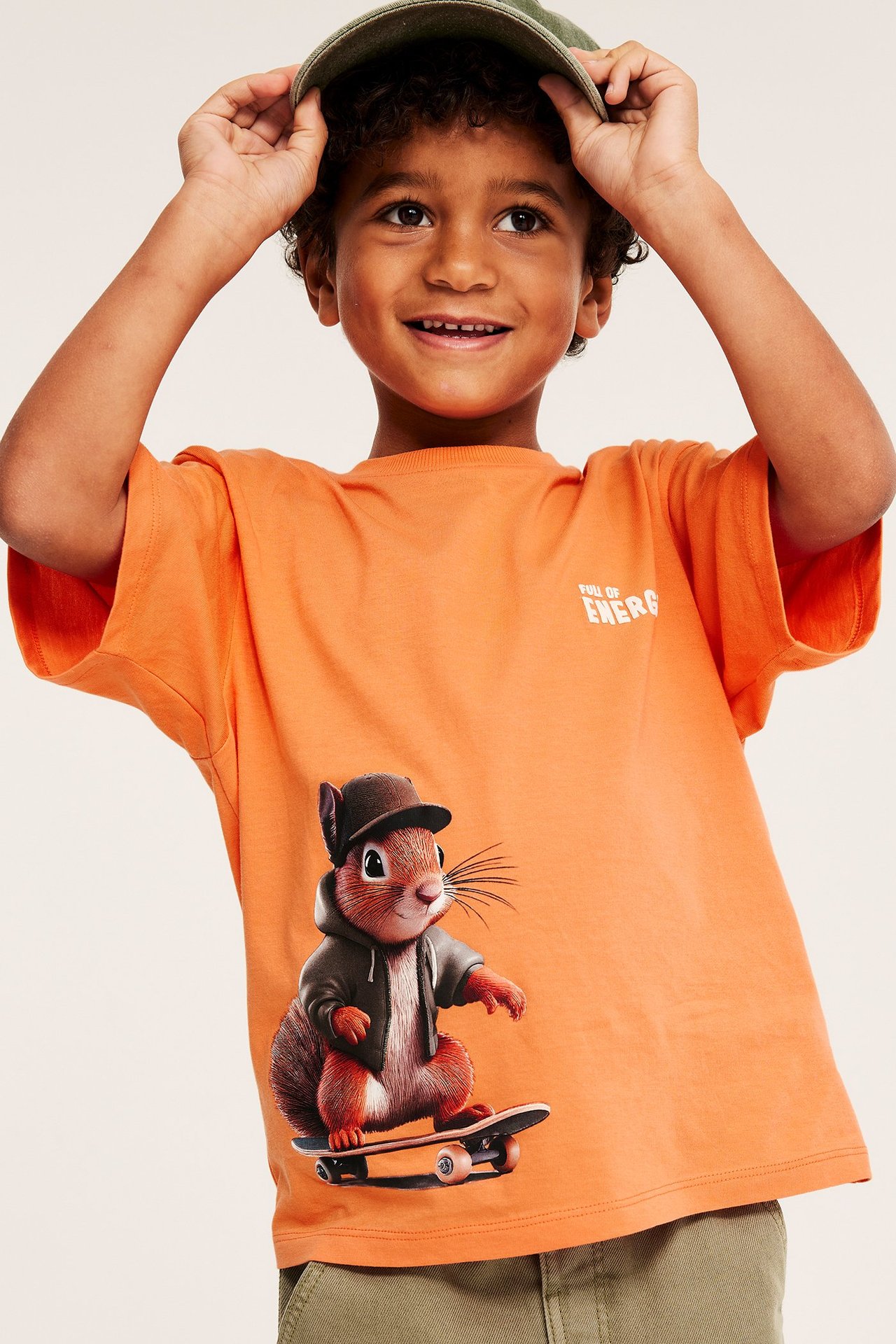 Etunäkymä: Lapsi oranssissa puuvilla-T-paidassa, jossa orava skeittilaudalla, ja vihreä lippis.