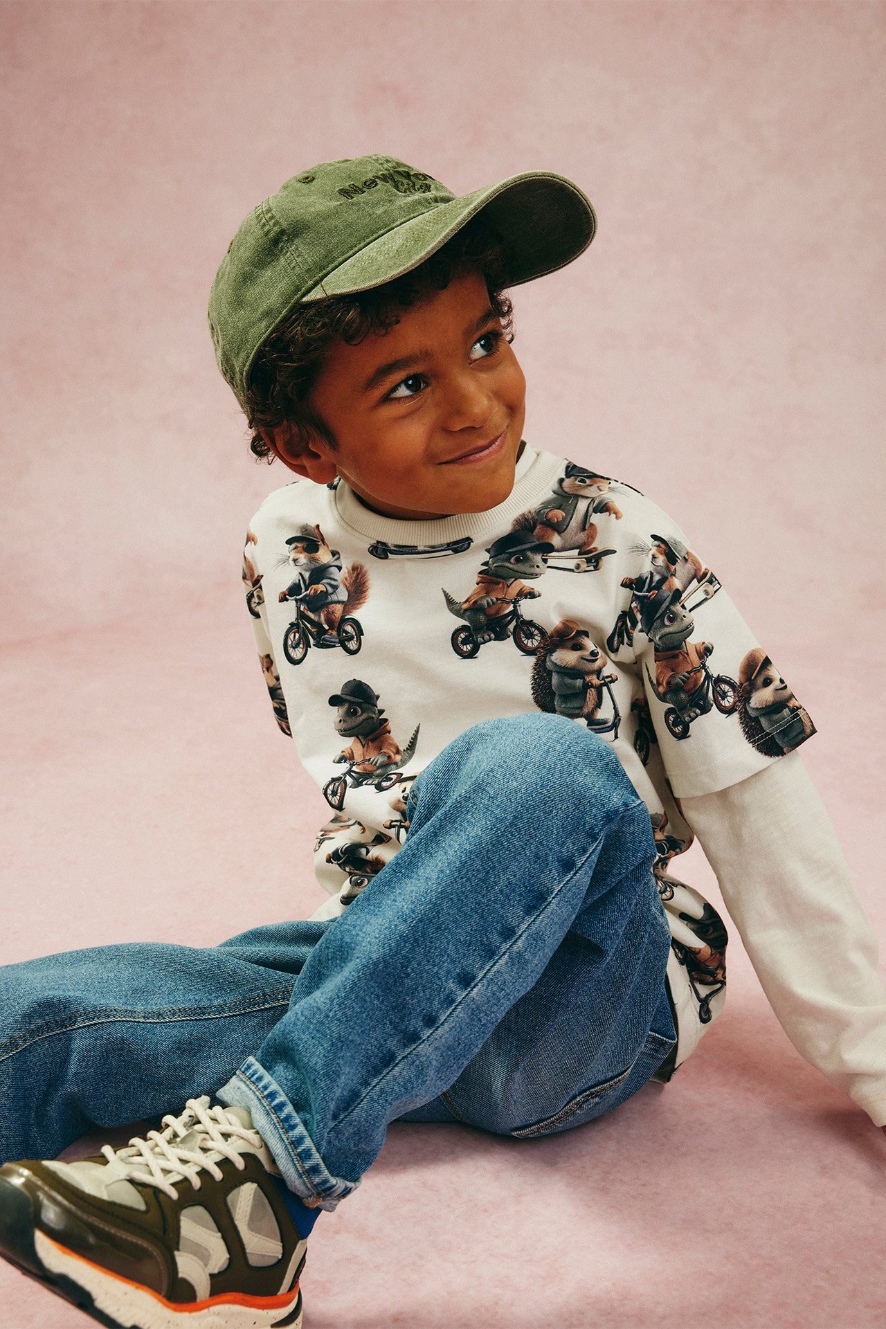 Frontvy av ett barn i offwhite bomulls t-shirt med djurmönster, blå jeans, grön keps och sneakers.