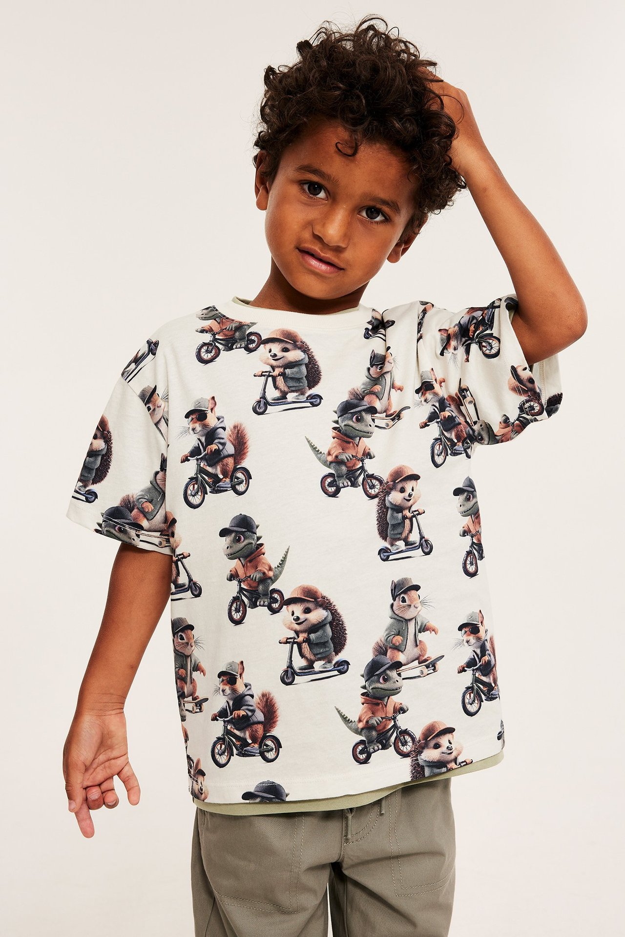 Edestäpäin kuvattu poikalapsi yllään luonnonvalkoinen puuvilla-t-paita eläinprintillä ja beiget housut.